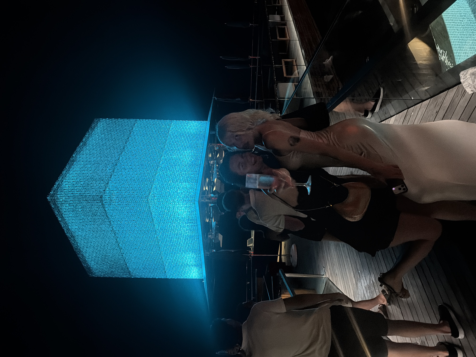 In the city of Kuta Selatan, Indonesia, at night, two women stand on a wooden deck in front of a large, cube-shaped structure illuminated with bright blue lights. The woman on the left, wearing a black dress, holds a champagne flute and a small tan handbag. The woman on the right, in a light-colored slip dress, has her arm around the other woman and is looking towards the camera. Behind them, several other people are visible, some in casual attire. The background features a dark sky and more wooden decking with seating areas. The overall scene suggests a nighttime gathering or event.