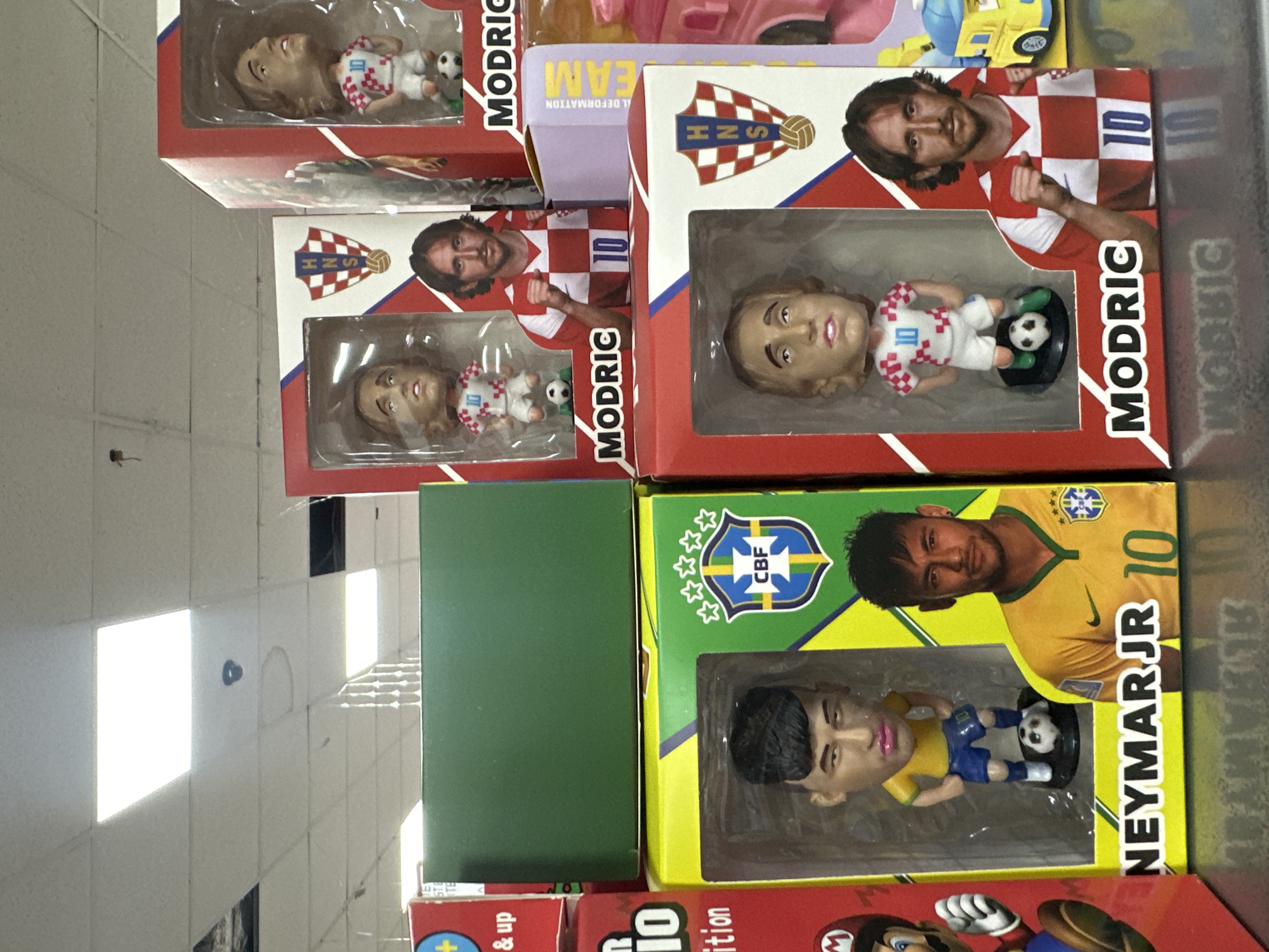 The image depicts a retail merchandise display in Budva, Montenegro, showcasing multiple boxed bobblehead figures of soccer players. In the lower-left foreground, a yellow and green box, prominently labeled "NEYMAR JR", contains a bobblehead figure of Neymar Jr. in a yellow and blue uniform with a soccer ball at his feet. The figure has distinct pink-colored lips. The box also features a photograph of Neymar Jr. and the CBF (Brazilian Football Confederation) logo with four stars.
To the right of the Neymar Jr. box, and extending into the mid-ground, are several red and white boxes, each labeled "MODRIC". These boxes contain bobblehead figures of Luka Modrić, uniformly dressed in a white and red checkered jersey with the number "10", standing on a black base with a soccer ball. Each "MODRIC" box also displays a photograph of Luka Modrić and the HNS (Croatian Football Federation) logo.
A dark green rectangular object is stacked directly above the "NEYMAR JR" box. Partially visible elements include a red box with an "M" logo, suggesting Mario merchandise, on the far left, and a purple box with "DEFORMATION" text alongside a yellow toy vehicle on the far right. The overhead environment features a white acoustic tile ceiling fitted with several rectangular fluorescent light panels providing illumination. A black, circular security camera is mounted on the ceiling in the upper-center portion of the image.