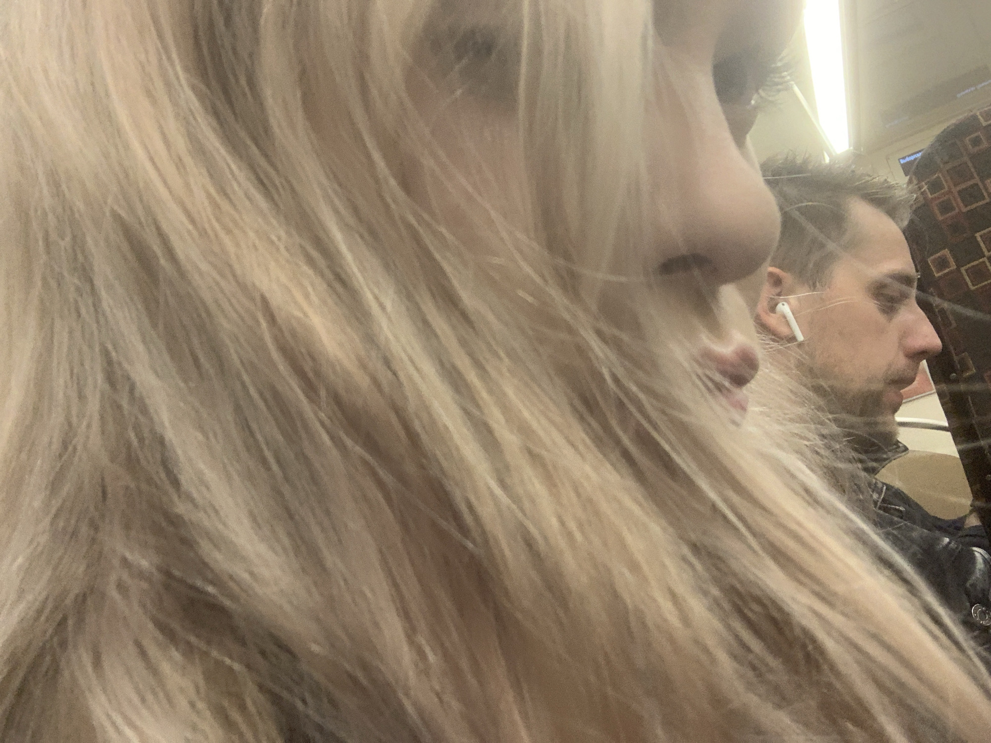 The image presents a close-up, candid view inside a public transport vehicle, likely a subway or train in Saint Petersburg, Russia. The foreground is dominated by the long, flowing blonde hair of a person, obscuring most of their face, though the side profile of their nose, mouth, and chin are partially visible on the left.
In the midground, to the right, a male passenger is seen in profile. He has short, dark hair, a beard, and is wearing a white wireless earbud in his right ear, suggesting he is listening to something. His gaze is directed downwards or forward, and his expression is neutral.
The setting features artificial overhead lighting and patterned upholstery on what appears to be a seat or wall in the background. Above the male passenger's head, some faint white Cyrillic text, possibly reading "Выберите" (meaning "Select" or "Choose"), is visible on a dark surface, consistent with Russian public transport signage. The image captures an ordinary moment of travel, with artificial light illuminating the interior. No specific time of day or weather conditions are discernible.