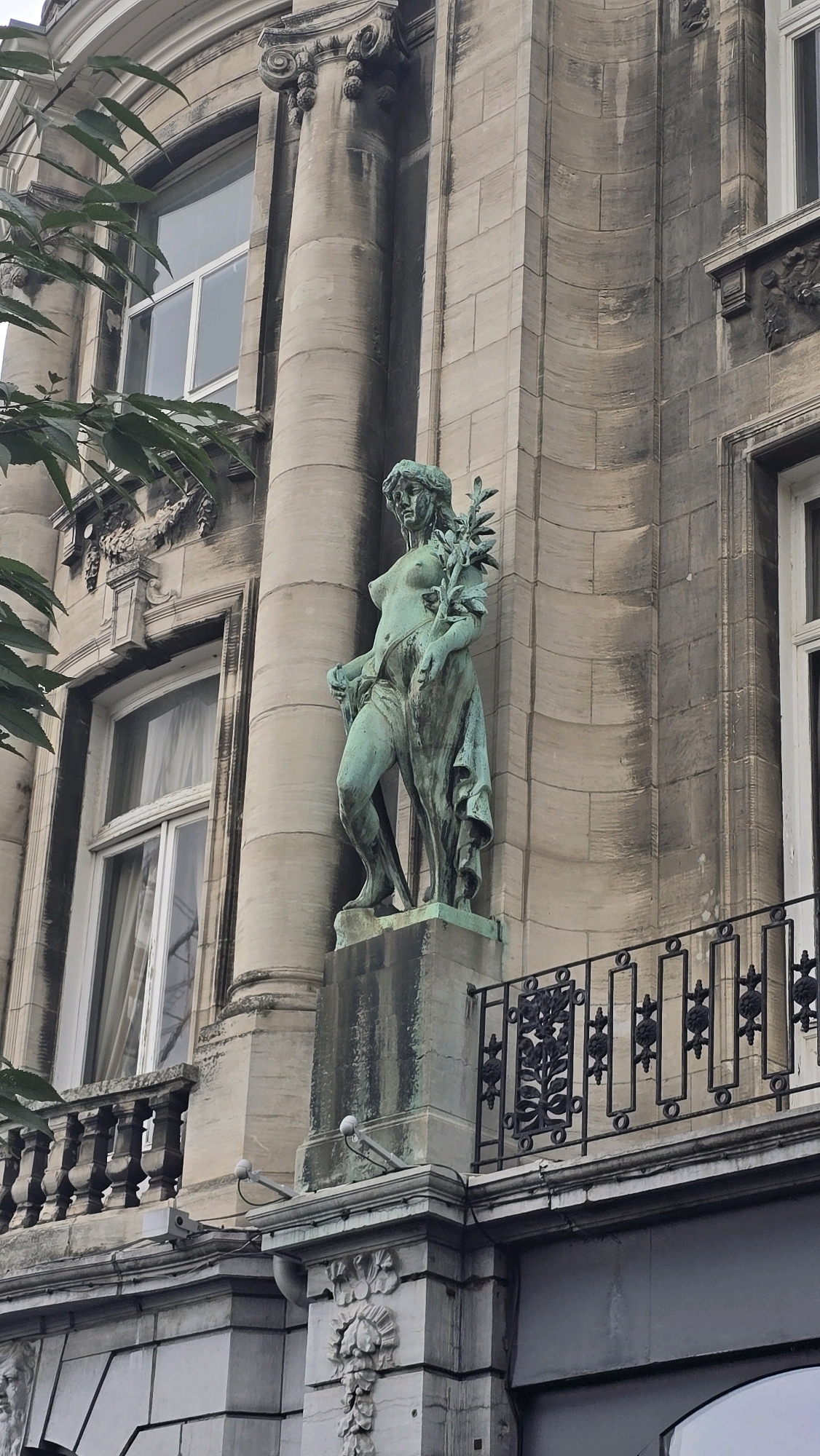 The image captures a close-up, eye-level view of a bronze statue of a female figure on the facade of a stone building in Antwerp, Belgium. The statue is depicted as standing and draped in a flowing garment, with one hand holding a branch with leaves. The building's architecture features classical elements, including large columns and ornate window frames. A black wrought-iron balcony railing runs across the lower portion of the image, adorned with decorative scrollwork. In the background, the facade exhibits textured stone blocks and appears somewhat weathered. The lighting suggests an overcast day. The statue and the building's details are the primary focus, with branches of a tree visible in the upper left corner.