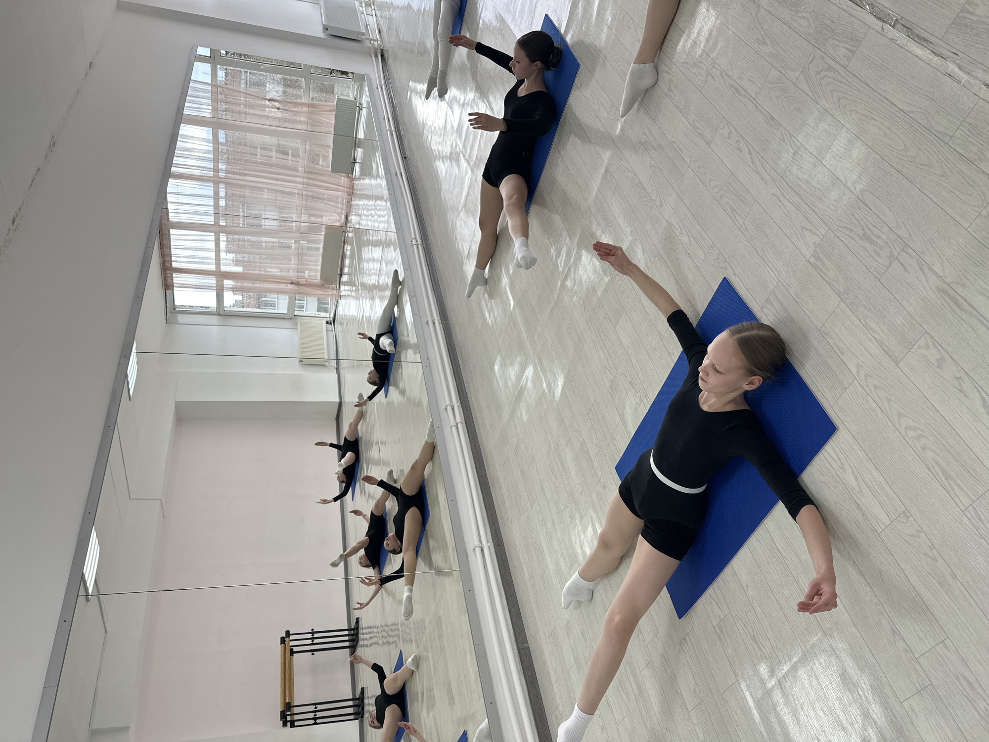 This image captures an indoor scene within a dance or gymnastics studio, likely located in Votkinsk, Russia. Several young girls, appearing to be pre-teens, are present, dressed in black athletic wear (leotards or tops with shorts) and white socks.
The main subject, positioned in the foreground, is a girl lying on her back on a blue yoga mat. Her arms are extended to her sides, and she has a calm, focused expression. Further back in the room, other girls are also visible, some lying on similar blue mats, suggesting they are resting or performing stretches.
A prominent feature of the studio is a large wall covered with floor-to-ceiling mirrors, which reflect the girls, the opposite wall, and ballet barres with metal frames and wooden rails. Large windows with white blinds are visible on an adjacent wall, allowing natural light into the room, indicating it is daytime. The studio has a light-colored, wood-patterned floor and white walls, with recessed light fixtures visible in the ceiling reflections. The overall scene depicts a training environment for physical activities like dance or gymnastics. There is no discernible text visible in the image.