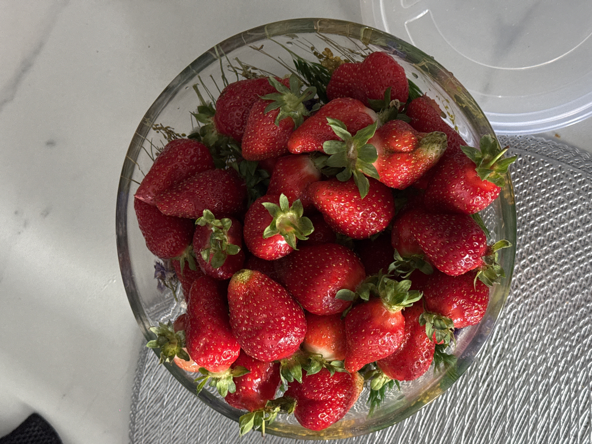 A top-down shot shows a glass bowl overflowing with fresh, red strawberries. The strawberries are piled high, with green stems visible amongst the fruit. Dried herbs are also visible inside the glass bowl. A white, marble-like surface is visible to the left of the bowl, and a textured, silver-colored placemat is on the right. The bowl's transparent plastic lid is seen in the background. This image provides no information related to Yerevan, Armenia.