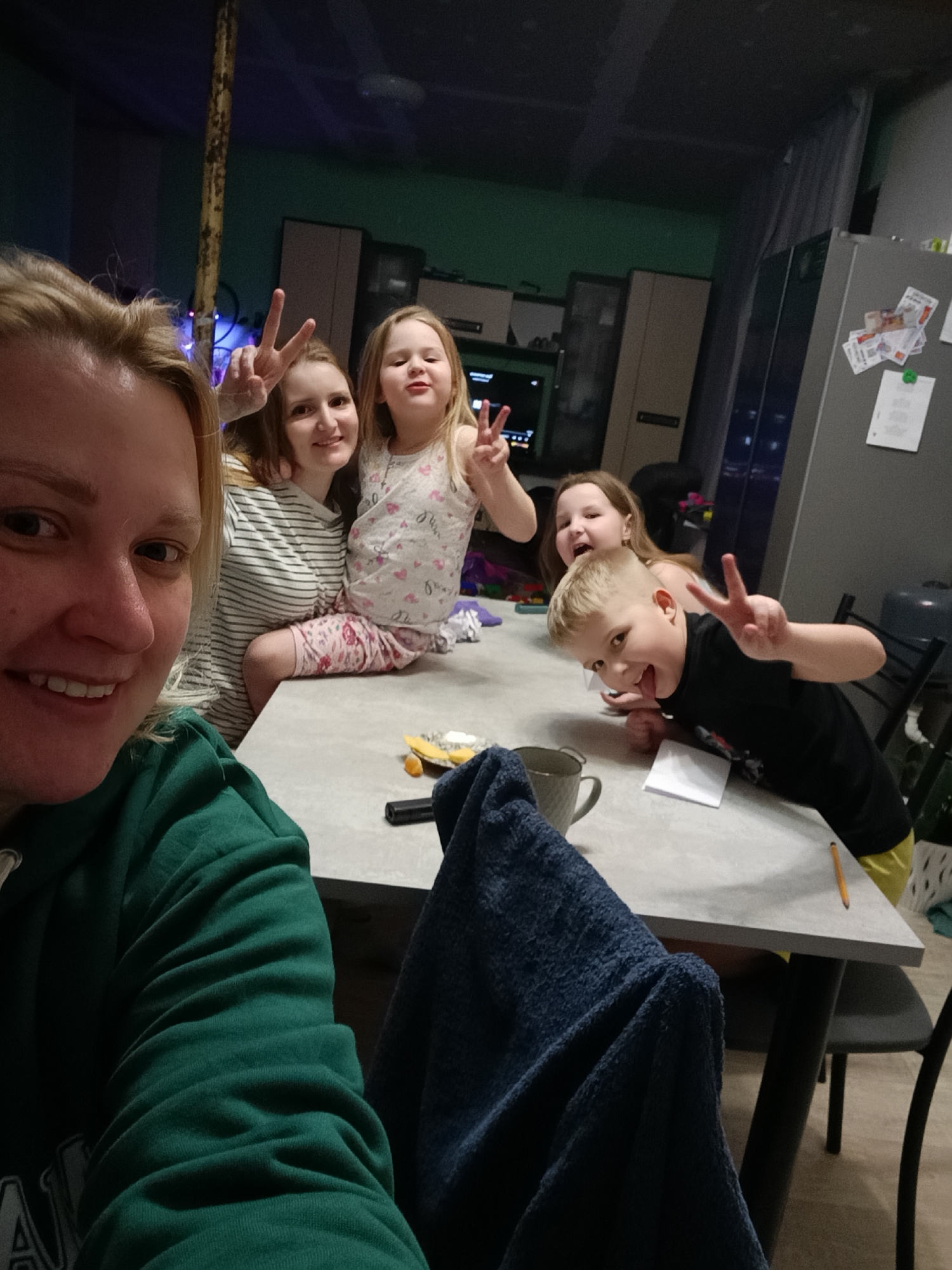 The photograph depicts a woman in a green sweatshirt taking a selfie with four children around a grey table. The subjects are making the "peace" sign with their fingers. On the table is a coffee mug, a black cylindrical object, a small white card, and some scattered items that appear to be food. A dark blue towel is draped over a chair. The background includes a refrigerator with papers attached, cabinets, and a television. The image was captured indoors in a residential area in Chaikovsky, Russia.