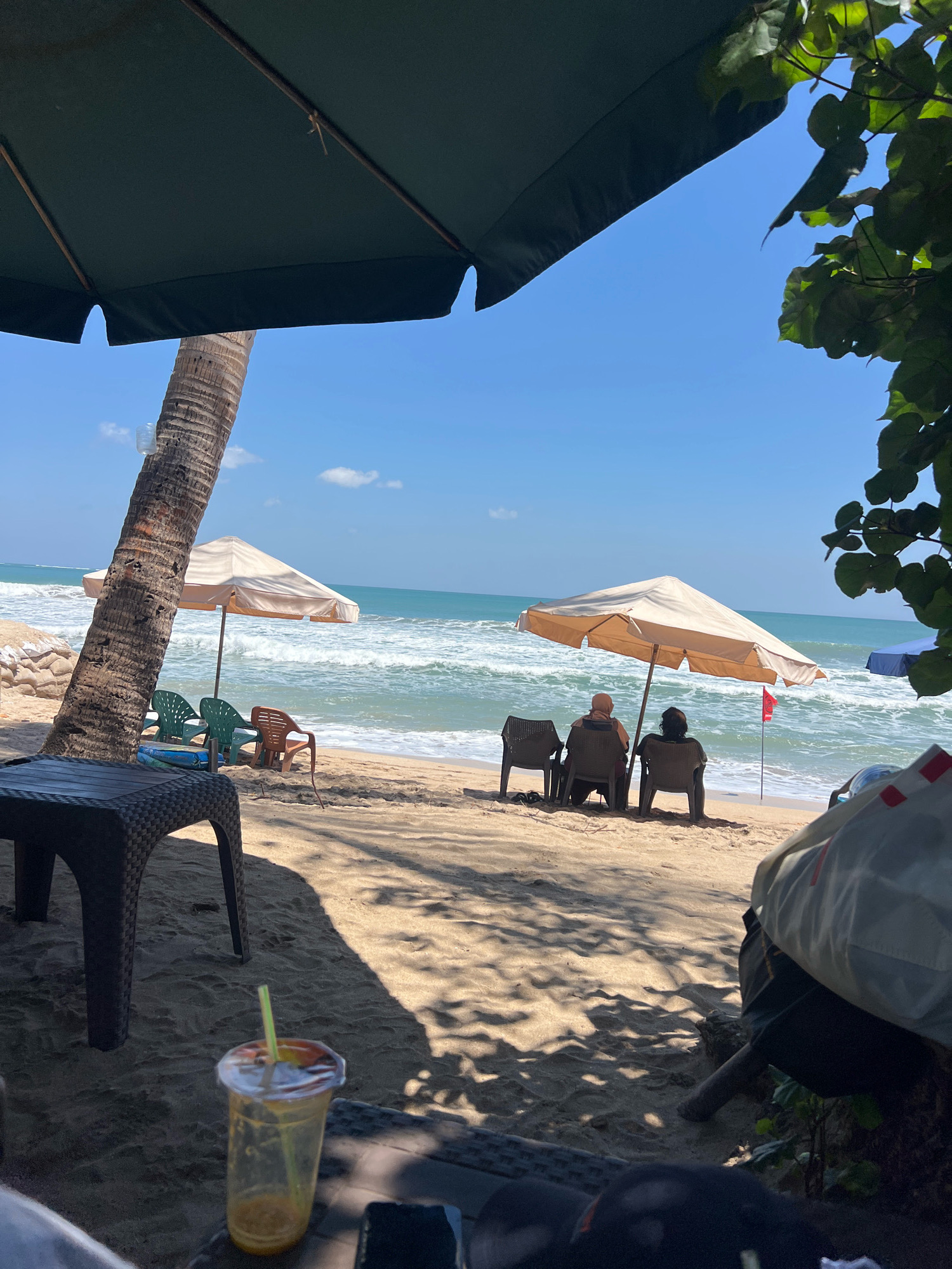 In Kuta, Indonesia, on a sunny day, two individuals are seated under a large umbrella on a sandy beach, facing the ocean. Waves are rolling onto the shore, and the water is a vibrant blue under a clear sky. In the foreground, a table and a partially consumed drink with a straw are visible. To the left, a palm tree stands tall. Further down the beach, another umbrella shades empty chairs. The scene suggests a relaxing day at the beach.