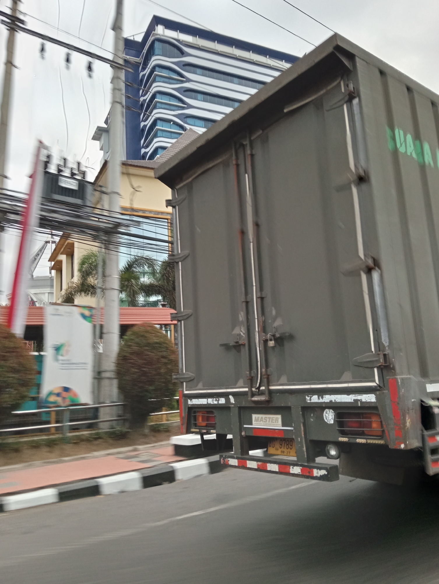An outdoor, eye-level, medium shot captures the rear of a dark green trailer truck in motion on a paved road. The truck's trailer fills the right half of the frame, its dark green surface showing wear and tear, with some white lettering faintly visible on its side. The rear of the trailer features various metal fixtures and latches, a red and white reflective bumper, and a yellow license plate displaying "AG 9789" with accompanying text below. To the left of the truck, a sidewalk with black and white striped curbing is visible, bordering a patch of grass and a short hedge. Behind the sidewalk, a white banner with colorful graphics and text is positioned between two small, rounded bushes. In the background, a multi-story building with a modern, wavy architectural design and blue-tinted windows stands tall, partially obscured by utility poles and a network of wires. A portion of a yellow building is also visible below and to the left of the modern structure. The sky above is overcast, suggesting a cloudy day. The motion blur indicates the photograph was taken from a moving vehicle.