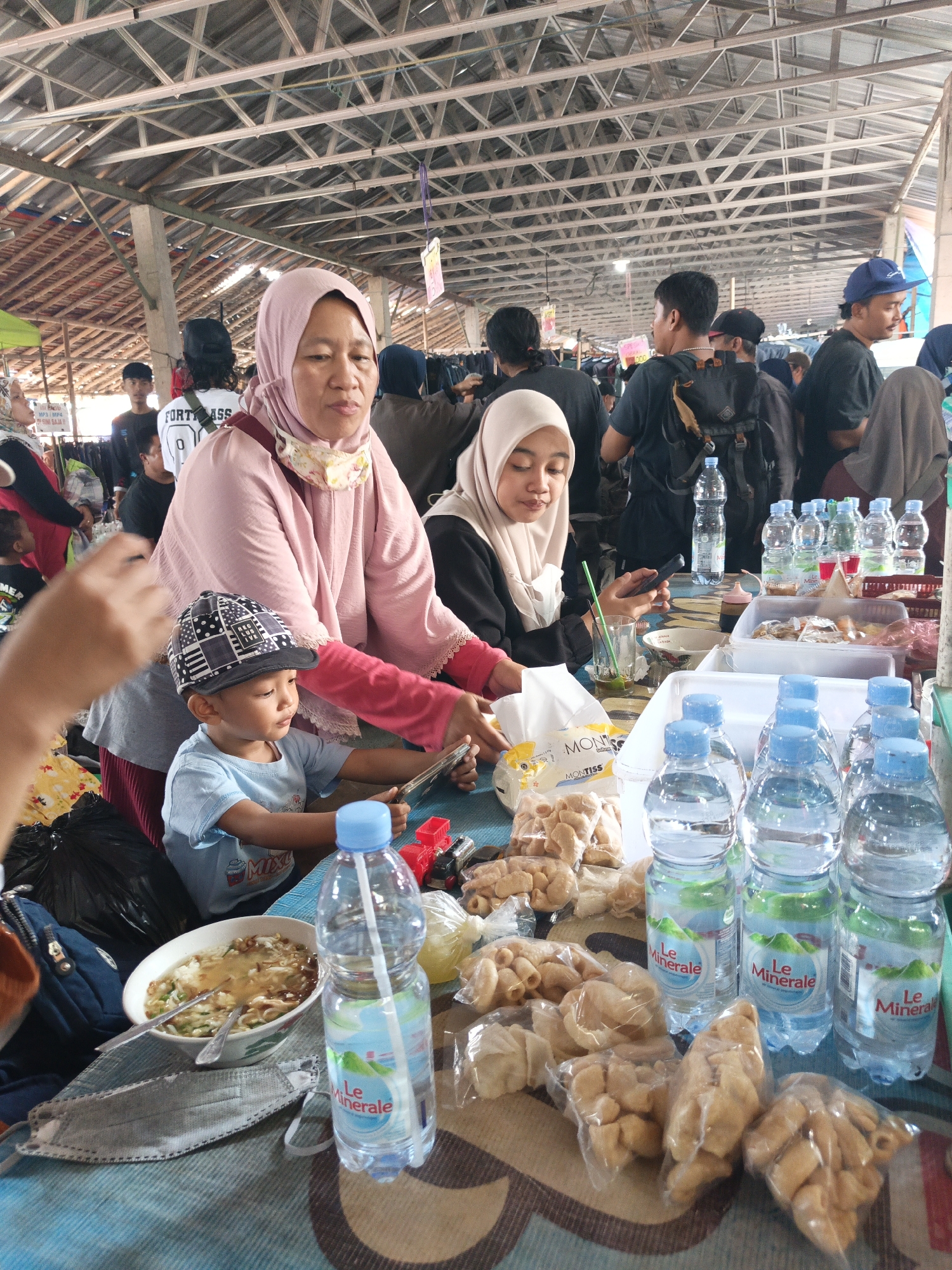 This image depicts a busy outdoor market scene, possibly in Jatinom, Indonesia, under a corrugated metal roof supported by a wooden structure. In the foreground, a young boy wearing a cap sits at a table, looking at a mobile phone. Next to him, a woman wearing a pink hijab and a patterned cloth around her neck is also at the table. The table is covered with a blue and white patterned cloth and is laden with food items, including a bowl of soup with noodles, several bottles of "Le Minerale" bottled water, and bags of fried snacks. To the left, a person's hand is visible, suggesting they might be holding the phone the boy is looking at. In the background, other individuals are milling about, some wearing head coverings, indicative of a public gathering. The overall atmosphere suggests a lively market environment.