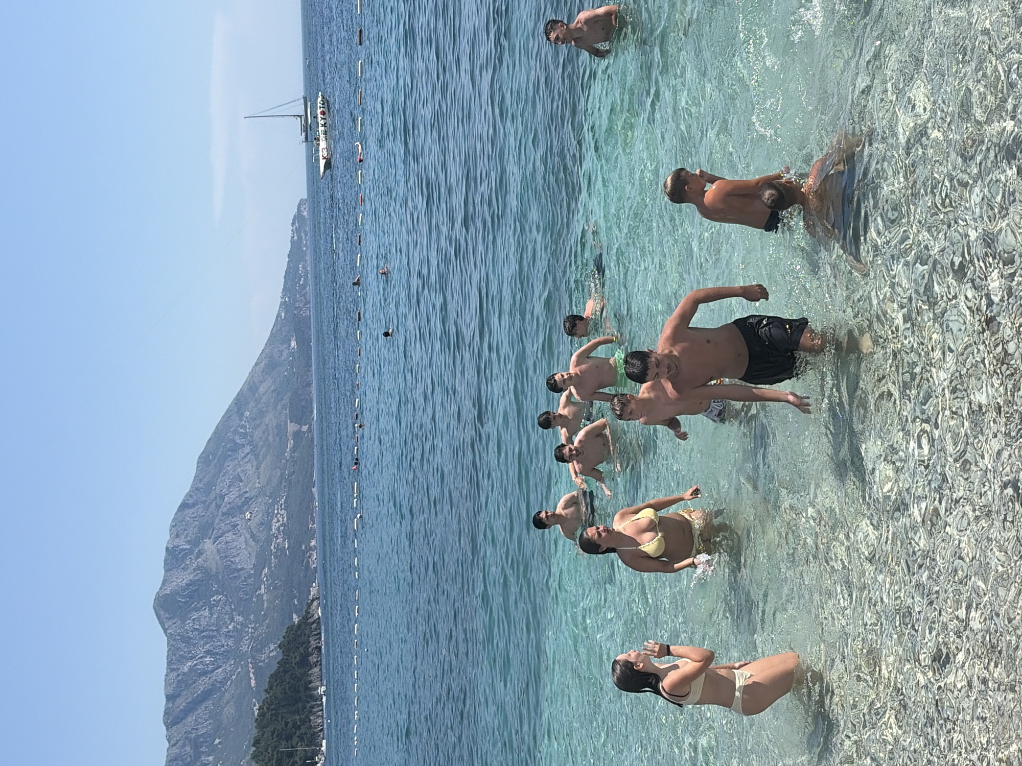 In Budva, Montenegro, a group of people are enjoying a sunny day at the beach. The turquoise waters of the Adriatic Sea are clear, revealing the pebble-strewn seabed. Several individuals, primarily young men and women, are wading and swimming in the water. One woman in a yellow bikini stands near the shore, while a young woman in a beige bikini is to her left, facing away from the camera and appearing to talk. A man in black shorts is walking towards the shore, emerging from the water. Other individuals are scattered throughout the water, some swimming, others standing. In the background, a majestic mountain range rises, covered in lush green vegetation. A sailboat with a white hull and mast is anchored in the distance, with other boats visible further out at sea. A rope with buoys stretches across the water, demarcating a swimming area. The sky is a clear, bright blue, indicating a pleasant summer day.