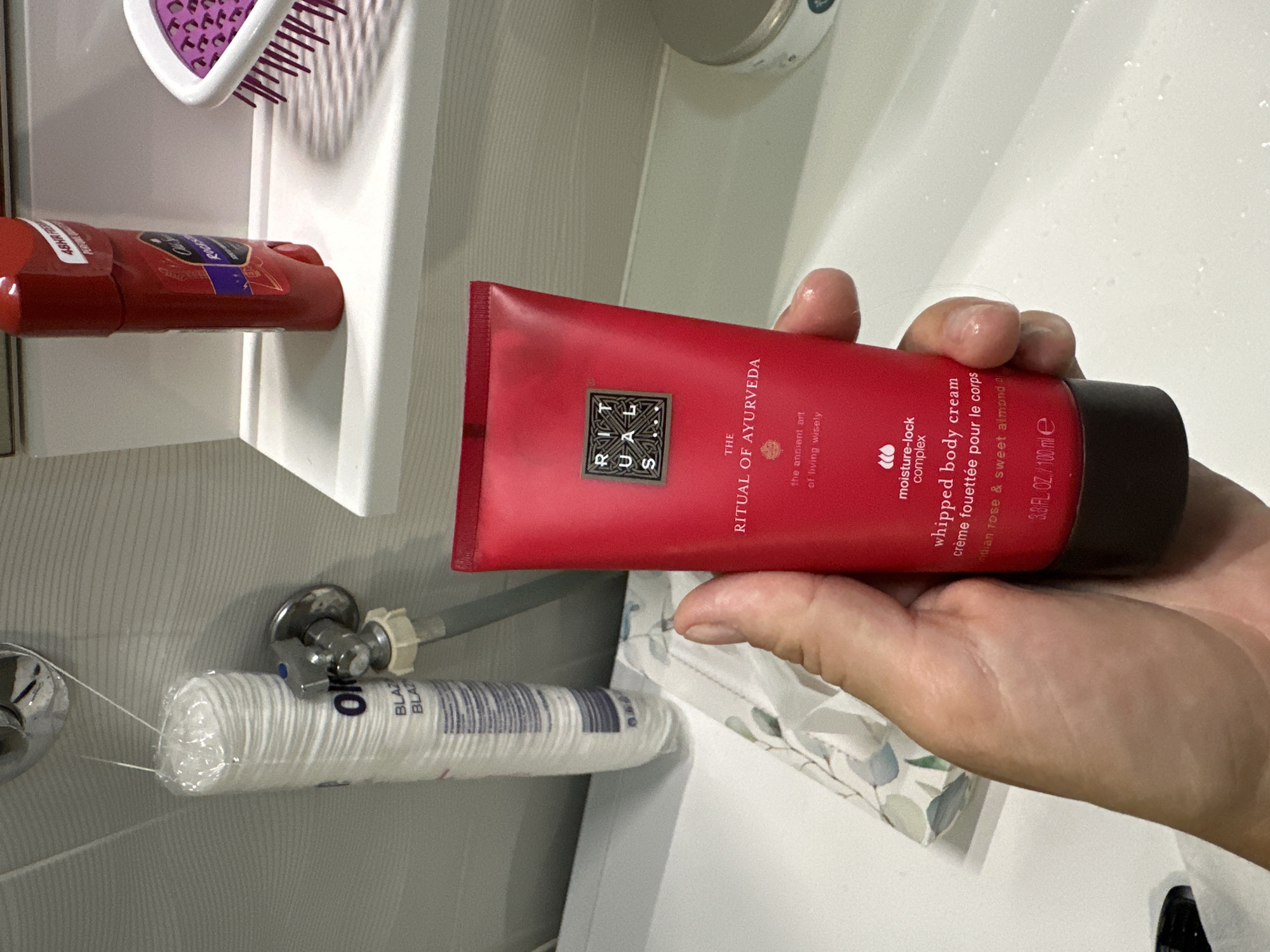 A hand holds a bright red tube of "The Ritual of Ayurveda" whipped body cream in a bathroom. The background features a white sink, a white shelf with a red stick deodorant and a purple-bristled brush, and a roll of cotton pads. The scene is indoors and appears to be a private residence bathroom. The location is not specified as Boreti, Montenegro in the image.