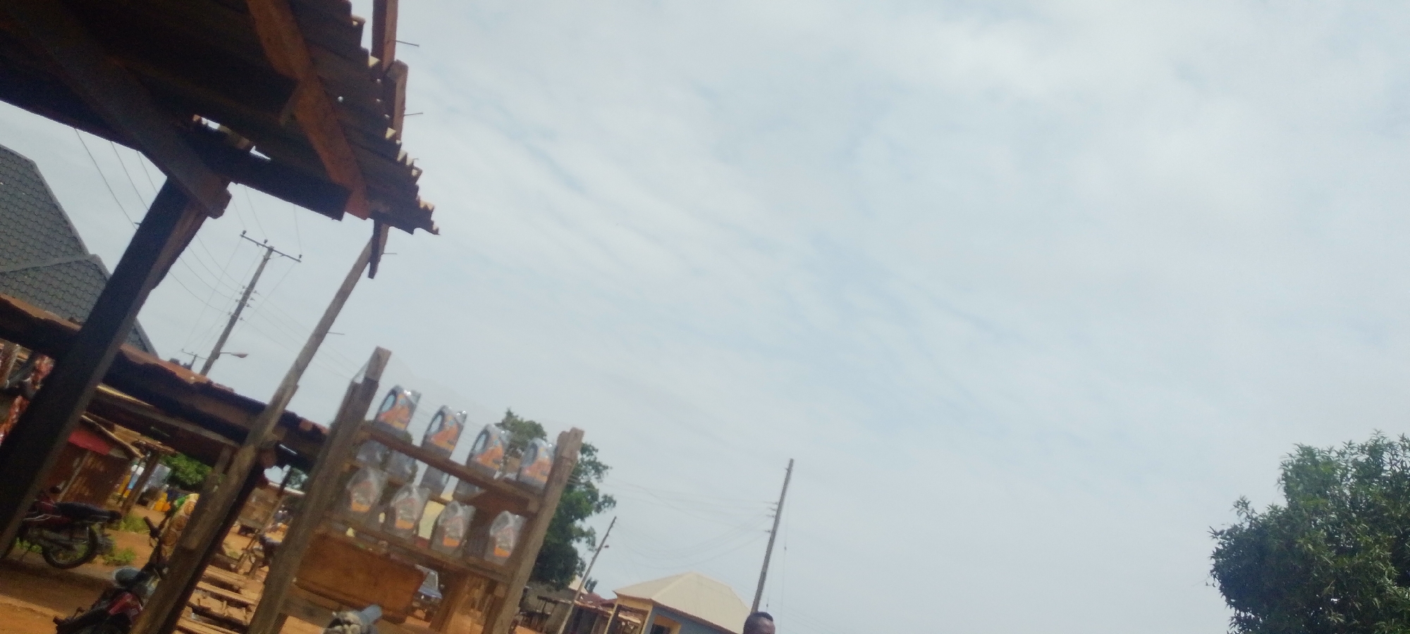 The image depicts an outdoor scene in what appears to be a market or roadside stall area in Baissa, Nigeria. In the foreground, a wooden structure with a corrugated roof is visible, with wooden beams supporting it. To the left, part of a house with a dark, tiled roof can be seen. Further left, a red motorcycle is parked.
On a wooden shelf in the mid-ground, several items are displayed, possibly bottles or containers, some of which appear to be wrapped in plastic. These are arranged in rows on the shelves. In the background, a utility pole with wires is visible, suggesting the presence of electricity infrastructure. A few other structures, possibly shops or homes, are also present in the distance, along with trees. The sky is overcast, indicating cloudy weather. The overall lighting suggests it is daytime, likely late morning or afternoon. There is no visible text in the image. The scene appears to be a typical daily life setting within a community in Nigeria.