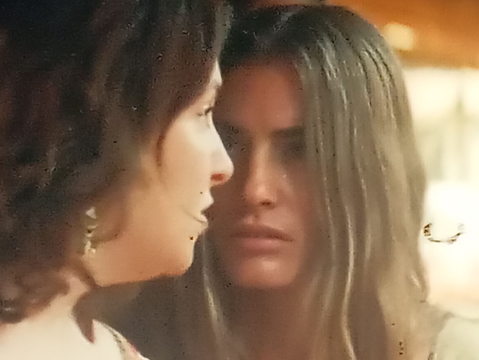 This is a close-up, slightly blurry photograph featuring two women. The woman on the left is shown in profile, looking towards the right. She has dark, wavy hair and is wearing earrings. Her face is soft and her expression is thoughtful. The woman on the right is facing forward, her gaze directed downwards and slightly away from the camera. She has long, light brown hair and her expression appears somber or melancholic.
The lighting in the image is warm and appears to be artificial, possibly from an indoor setting. The background is out of focus, suggesting a shallow depth of field, and is rendered in soft, warm tones of orange and yellow, hinting at an interior with ambient lighting. The overall impression is intimate, perhaps a moment of quiet contemplation or emotional connection between the two individuals. The image has a slightly grainy texture and some visible specks, possibly dust or imperfections on a lens or surface. No text is visible in the image. The context provided places this scene in Sofiivska Borshchahivka, Ukraine.