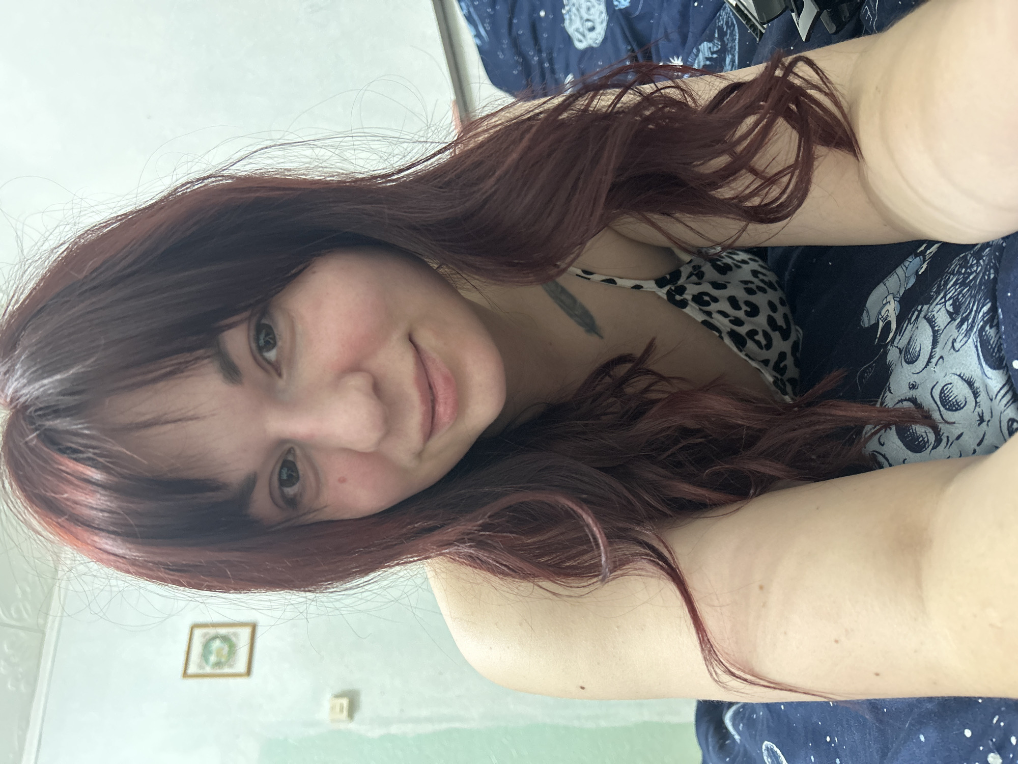 A selfie shows a woman with auburn hair and dark bangs. She is smiling slightly at the camera. She has fair skin and visible eyelashes. Her top has a black and white animal print. A tattoo is visible on her collarbone. She is lying on a dark blue bedspread with white celestial designs. A corner of what appears to be a desk is visible behind her. The wall is light green with a framed picture. There is no indication of the location being in the city of Фоки, Russia.