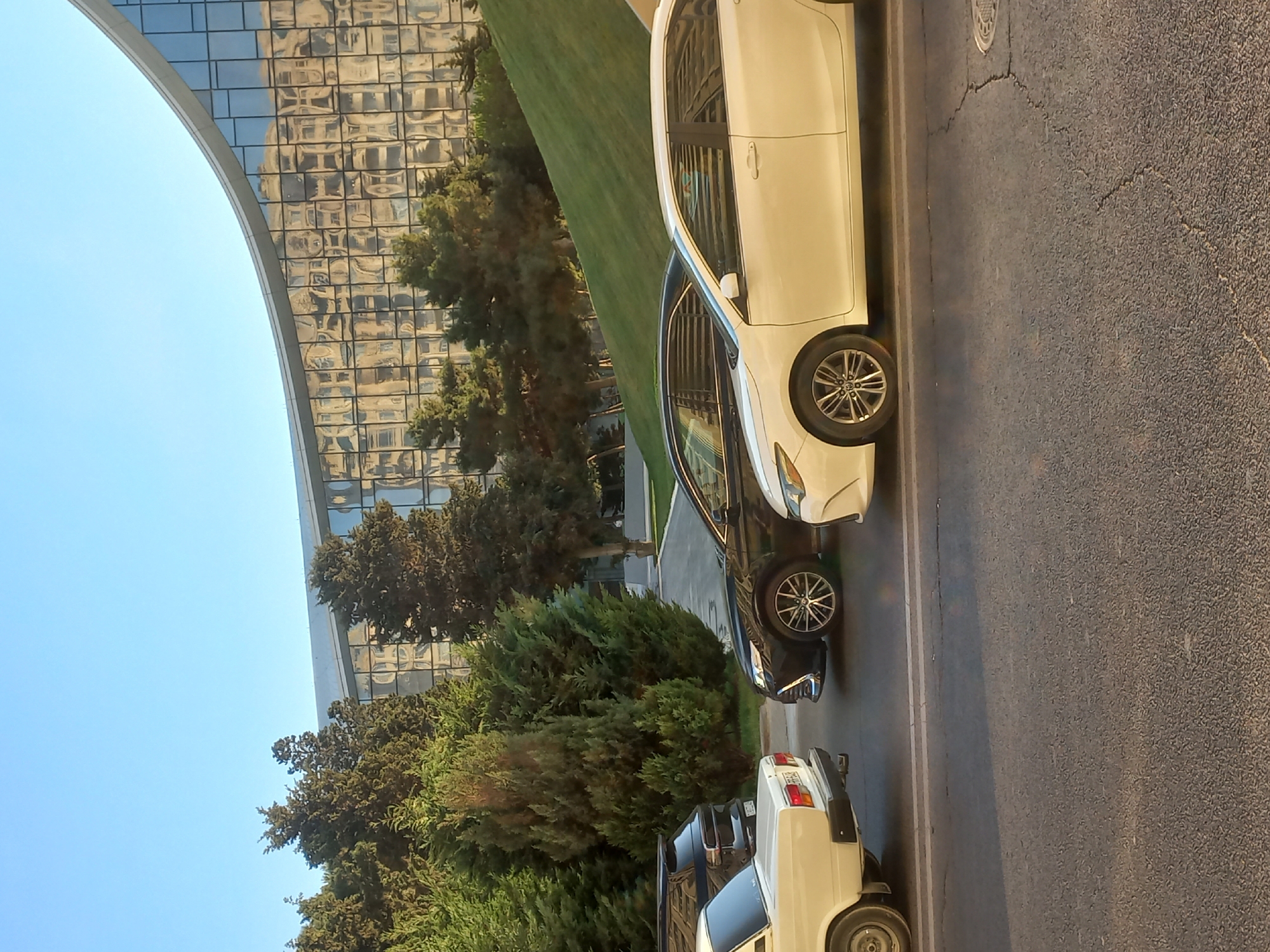 In Baku, Azerbaijan, a street scene unfolds with a modern, curvilinear building reflecting the clear blue sky and surrounding trees. Several cars are parked along the roadside, including a white sedan, a black sedan, and a black SUV. The white sedan in the foreground, possibly a Lada, is parked parallel to the street, its rear bumper facing the camera. Next to it, a black sedan, likely a Toyota Camry, is also parked. A white sedan, possibly a Toyota Camry, is visible in the background on the right. The street is paved with asphalt, showing some cracks. A grassy slope rises towards the building, dotted with evergreen trees. The lighting suggests it is late afternoon or early evening, with the sun casting warm tones on the scene. There are no people visible in the image. The overall impression is one of urban modernity and natural elements harmoniously coexisting.