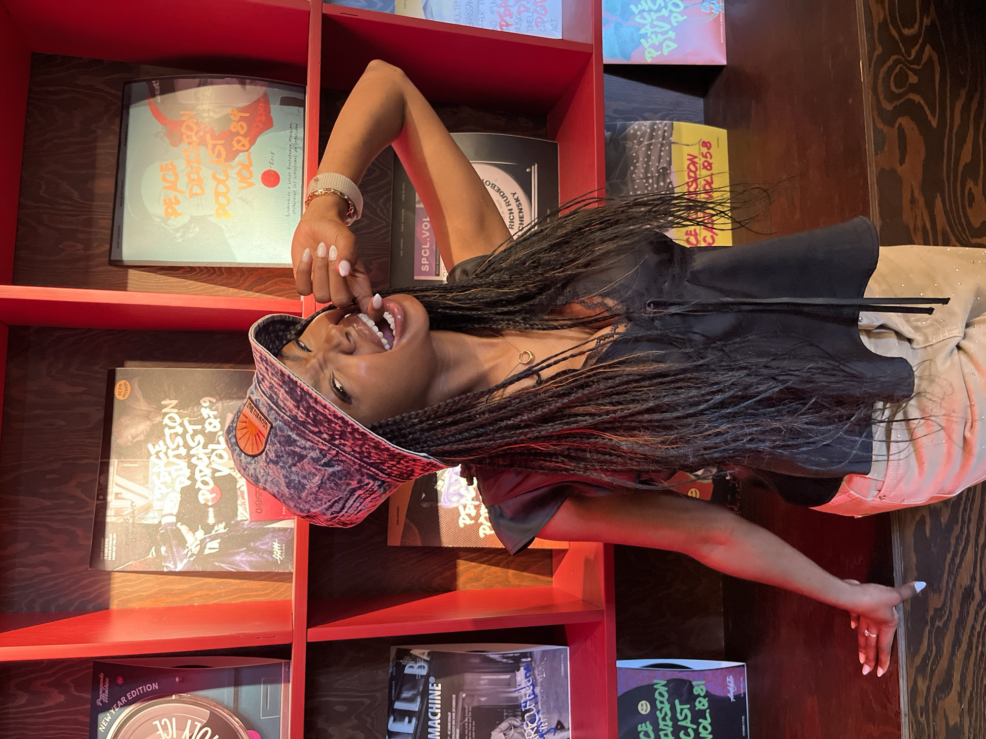 A young Black woman with long braided hair, wearing a denim bucket hat and a black top, poses in front of a red shelf filled with vinyl records. She is leaning on a wooden counter, with her right arm raised and her fingers near her mouth. Her left hand rests on the counter. The records on the shelf are mostly covered, but some titles like "Peace Division Podcast Vol. 079" and "Peace Division Podcast Vol. 084" are visible. The overall setting appears to be a record store or a similar establishment. The location is specified as Kazan, Russia.
