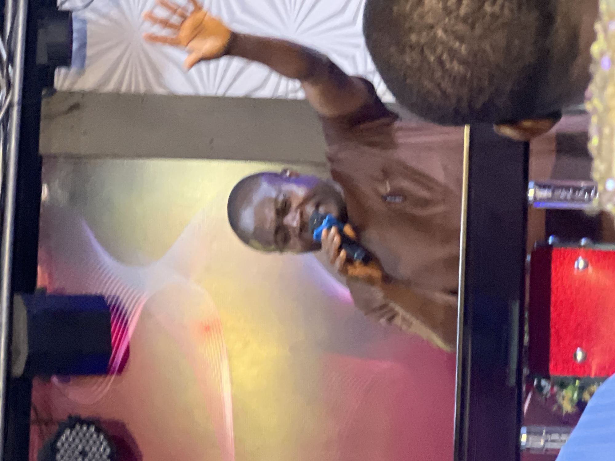 In Nkpor, Nigeria, a middle-aged Black man, wearing a brown t-shirt, is captured mid-action on a stage. He holds a blue microphone to his lips, suggesting he is speaking or singing. His right arm is raised, hand open, in a gesture that could signify praise, greeting, or emphasis. The background is a vibrant, abstract display of colored lights and patterns, possibly from stage lighting or a decorative backdrop, with a white, textured panel on the right. In the foreground, a dark surface, likely a table or console, partially obscures the view of a bright red object below. The scene appears to be a performance or an event taking place indoors, with other audience members present, one of whom's head is visible in the lower right corner.