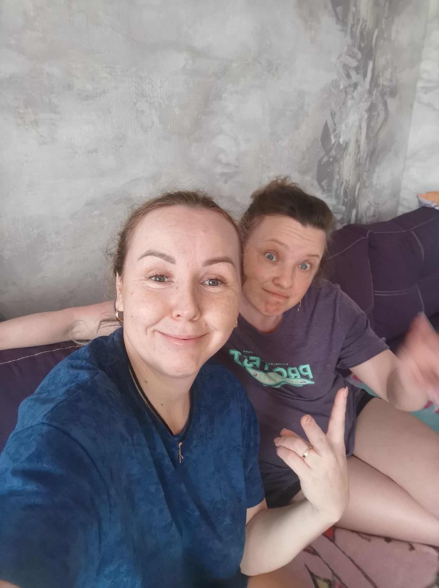 Two women are pictured in a selfie. The woman on the left is in the foreground, smiling and looking at the camera. She is wearing a blue t-shirt. The woman on the right is in the background, looking at the camera with a surprised expression and her mouth open. She is wearing a purple t-shirt with a graphic design. They are both sitting on a couch in front of a wall with a textured gray finish. The background wall has a dark, abstract pattern on the right side. The couch is covered in a dark fabric. The lighting is natural, suggesting it is daytime indoors. The image appears to be taken in an apartment or home setting.