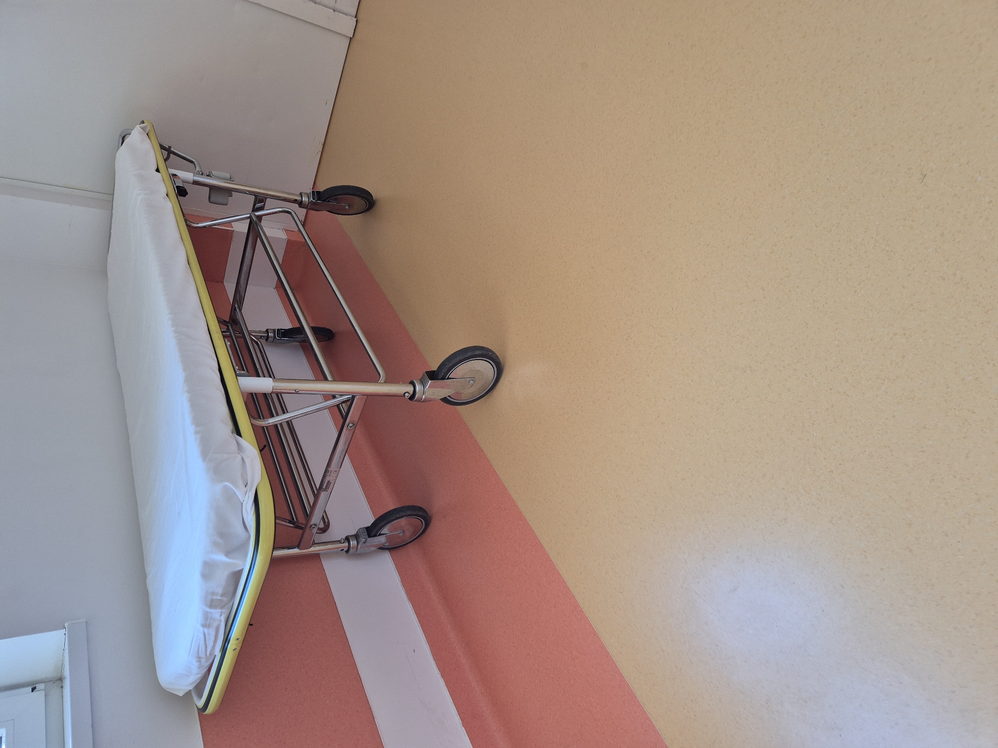 In an indoor setting, a gurney with a white sheet is positioned diagonally on a floor that transitions from a peach color near the wall to a light tan color further into the room. The gurney has a yellow trim and a metal frame with four wheels. The wall behind the gurney is white, with a horizontal stripe of peach and white visible along the bottom portion. To the left of the gurney, a sliver of a window is visible. The scene is brightly lit, suggesting an institutional environment such as a hospital or clinic.