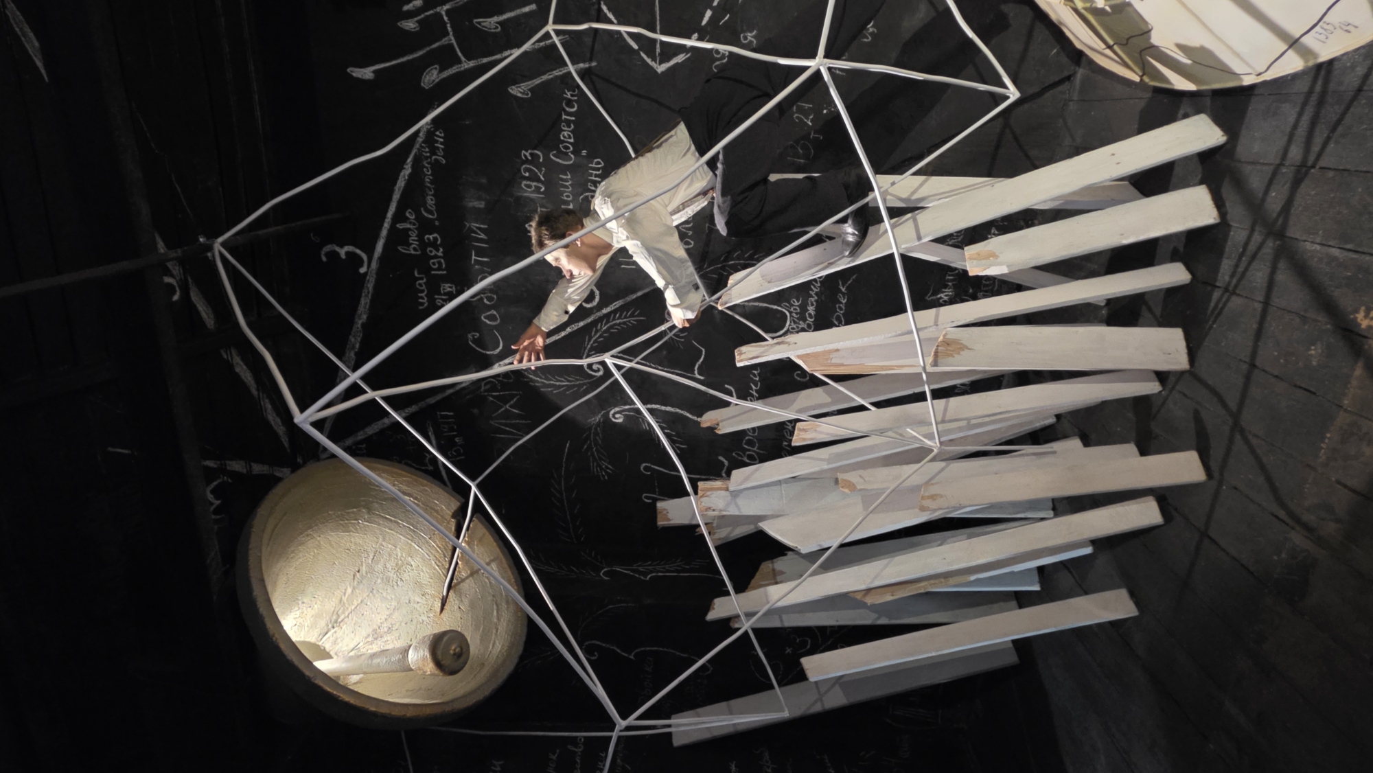 A tilted, low-angle shot shows a person in the center of a stage set, possibly in Saint Petersburg, Russia. The individual, dressed in a light-colored shirt and dark pants, is crouched and reaching out to a white geometric wireframe structure. The backdrop is a black wall covered in white chalk inscriptions. A lamp or lighting fixture is positioned to the left of the person, adding a glow to the scene. Wooden planks are arranged haphazardly around the wireframe structure.