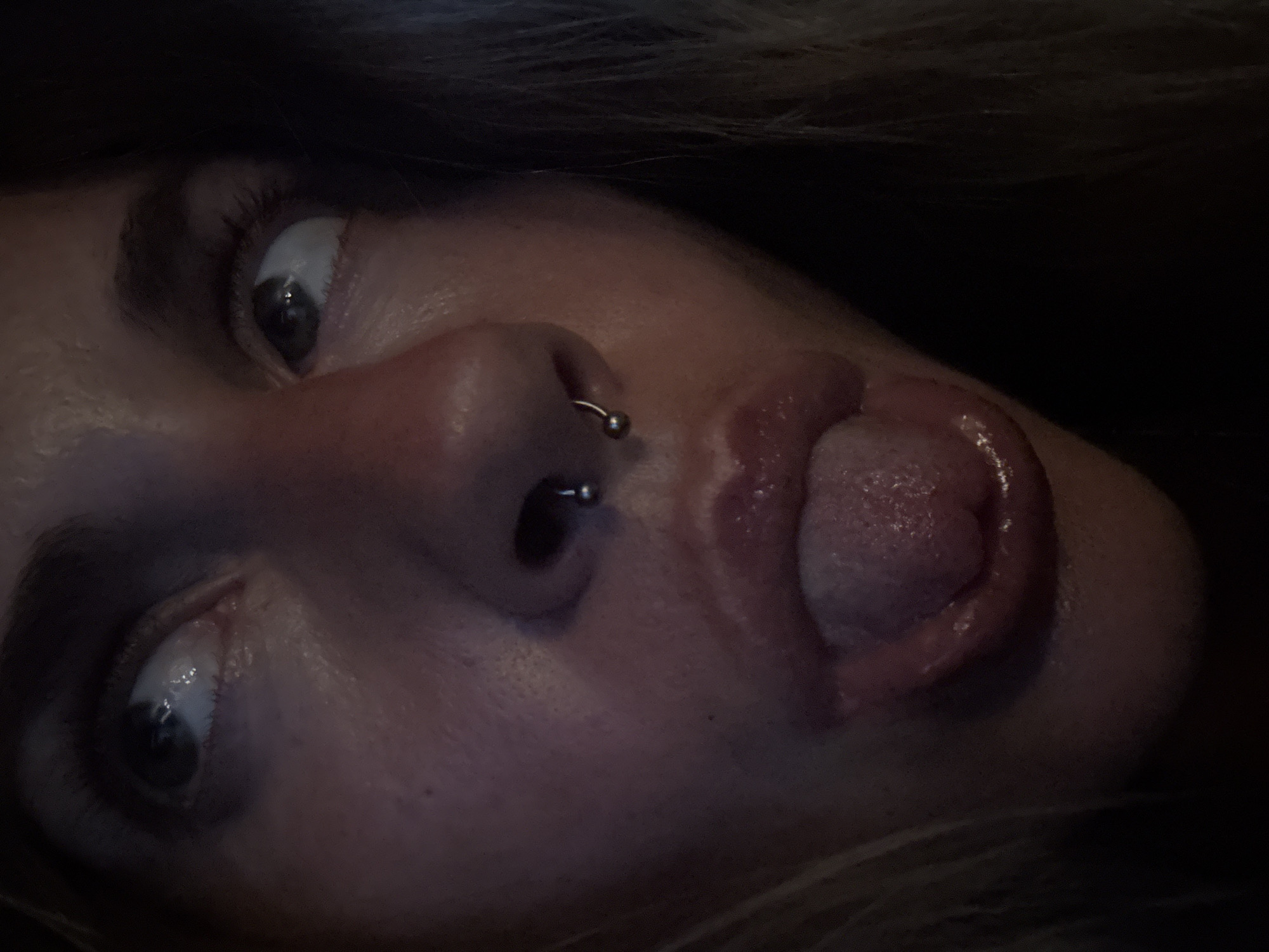 A close-up, overhead, natural light, indoor shot shows the face of a person with their tongue sticking out. The person's face is partially obscured by shadow. The person is looking upwards and to the right. The person has a septum piercing. The background is dark and out of focus.