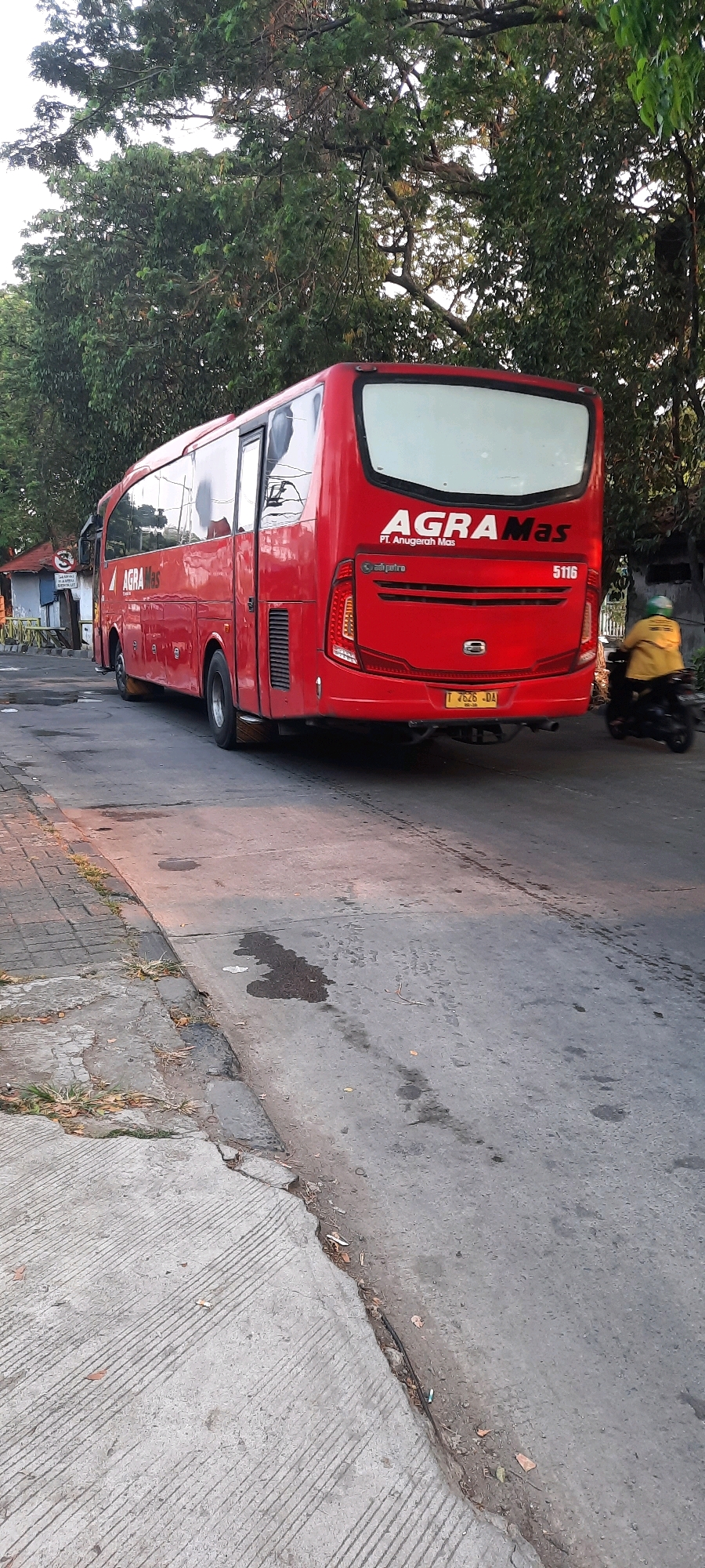 A red bus, identified as "AGRA Mas" with the text "PT. Anugerah Mas" below it, is parked on the right side of a street in East Jakarta, Indonesia. The bus is facing away from the viewer, showing its rear and right side. The number "5116" is visible on the back of the bus. A yellow motorcycle with a rider wearing a yellow jacket and green helmet is passing the bus on the right. The street is paved with asphalt and there is a concrete sidewalk on the left. Trees with green foliage line the background. The lighting suggests it is daytime, possibly late afternoon given the warm tones on the road.
