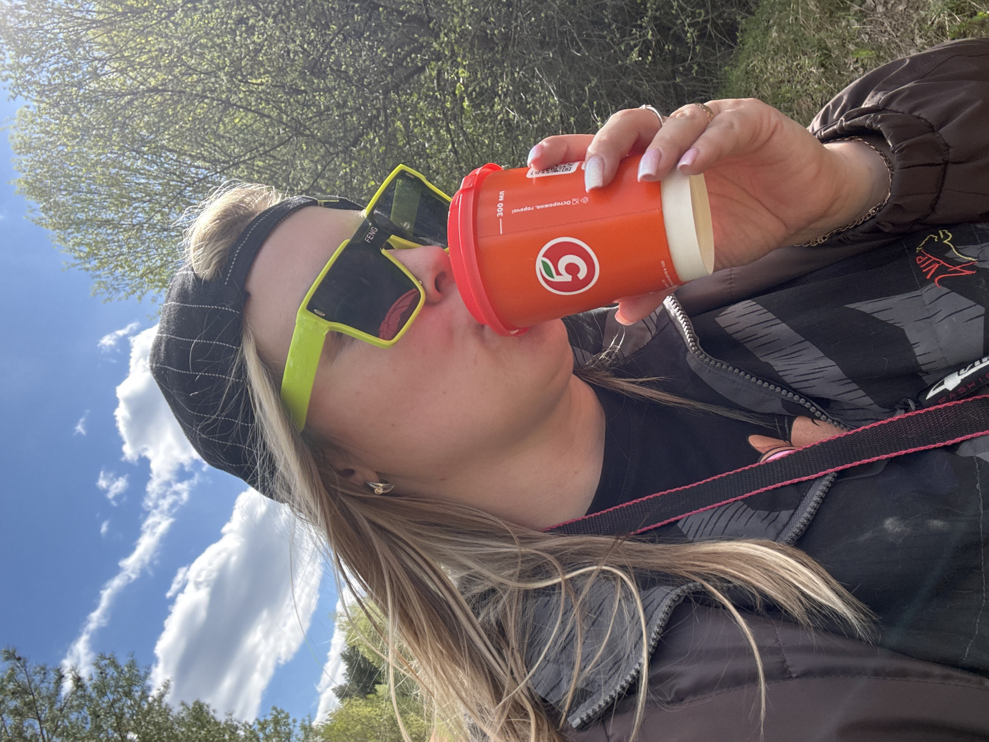 A woman drinks from an orange cup while wearing sunglasses. She is wearing a black and gray jacket with a black hat. The background includes trees and a blue sky with white clouds. The cup has the logo of a Russian grocery chain, indicating the location as likely Марковский, Russia.