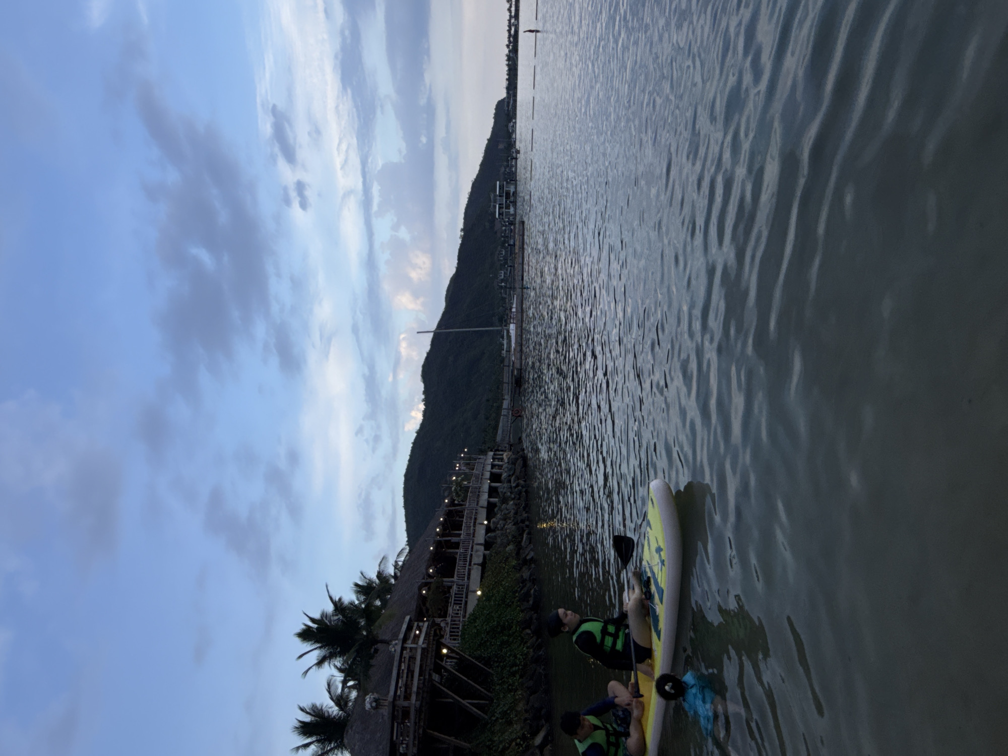 The image captures a twilight scene in Bac Nha Trang Ward, Vietnam. In the foreground, two individuals wearing life vests are seated on a yellow paddleboard in the water. One holds a paddle. The water's surface is rippled. Adjacent to the water is a shoreline with a building constructed from wood and thatch. The building is illuminated with lights. The background consists of a mountain range rising into a cloudy sky.