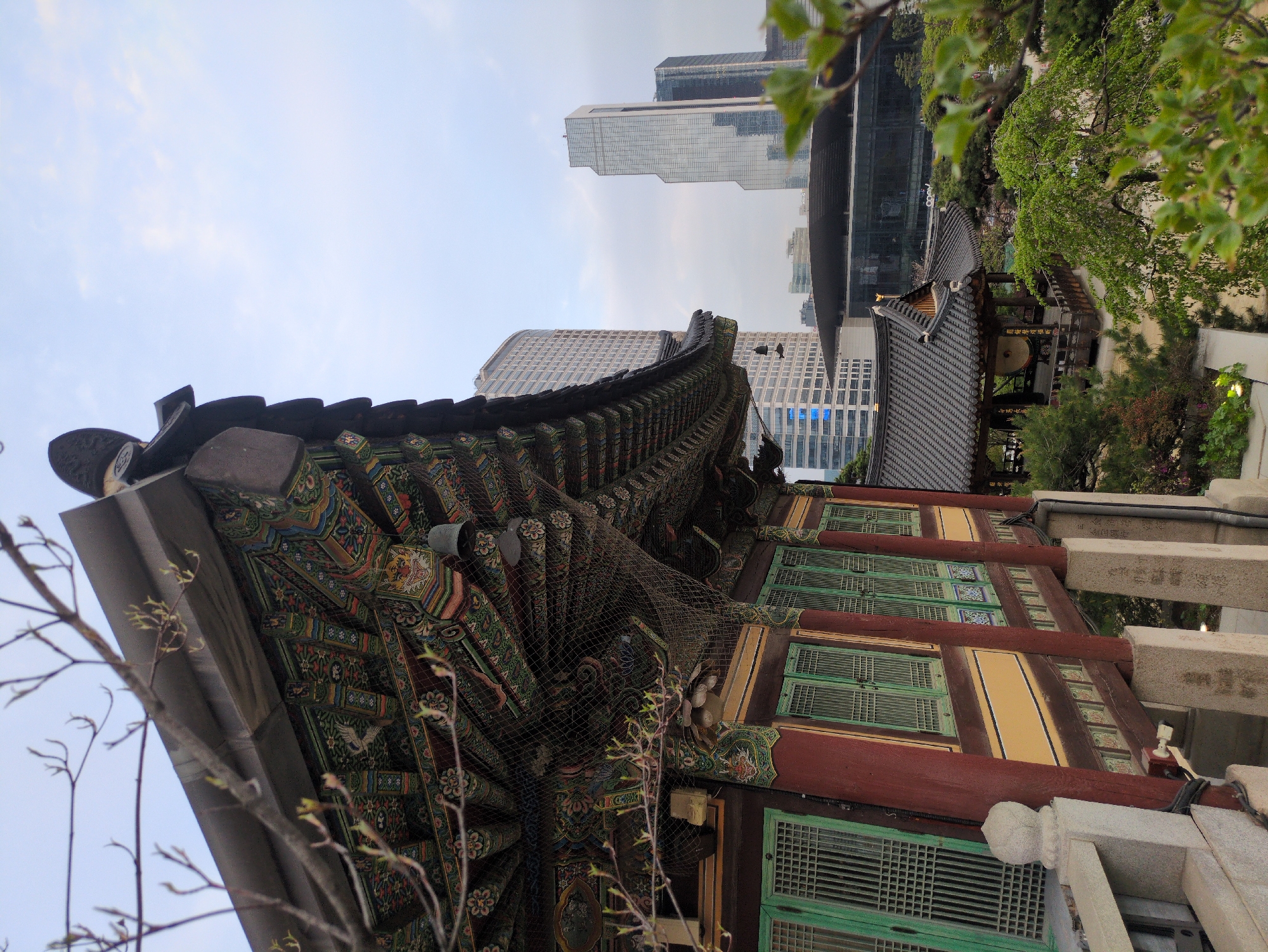 The image shows an eye-level, medium shot of the exterior of a traditional Korean building in Seoul, South Korea. The building is multi-colored, with intricate geometric patterns and traditional Korean architectural details. The walls are yellow, with red accents and green shutters. The roof is dark, with ornate eaves. A fine net is draped over the structure.
In the background, modern skyscrapers rise, highlighting the juxtaposition of the old and new in Seoul. A smaller, tiled-roof structure is visible to the right of the main building. Green trees surround the buildings.
The sky is overcast. There are bare tree branches in the foreground. The image suggests a peaceful scene within a bustling urban environment.