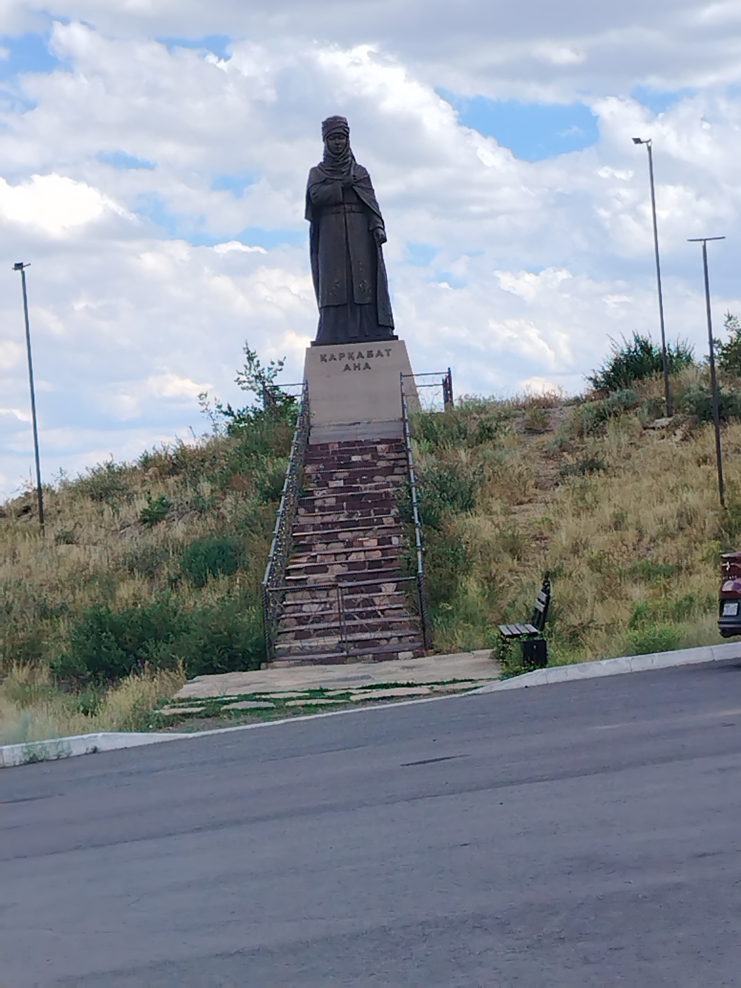 The image shows a statue of a woman dressed in traditional attire, standing on a pedestal. The pedestal has text inscribed on it. A set of brick stairs leads up to the pedestal, and a metal railing runs along the side of the stairs. The statue is situated on a grassy hill with scattered bushes and trees. In the background, there are two tall lampposts and a cloudy sky. To the right of the stairs, a wooden bench with a black trash can is visible. Further to the right, a portion of a vehicle and a wooden fence can be seen. The scene appears to be outdoors, possibly a park or a memorial site, in Qarqaraly, Kazakhstan. The lighting suggests it is daytime. The text on the pedestal reads "ҚАРҚАРАЛЫ АНА".