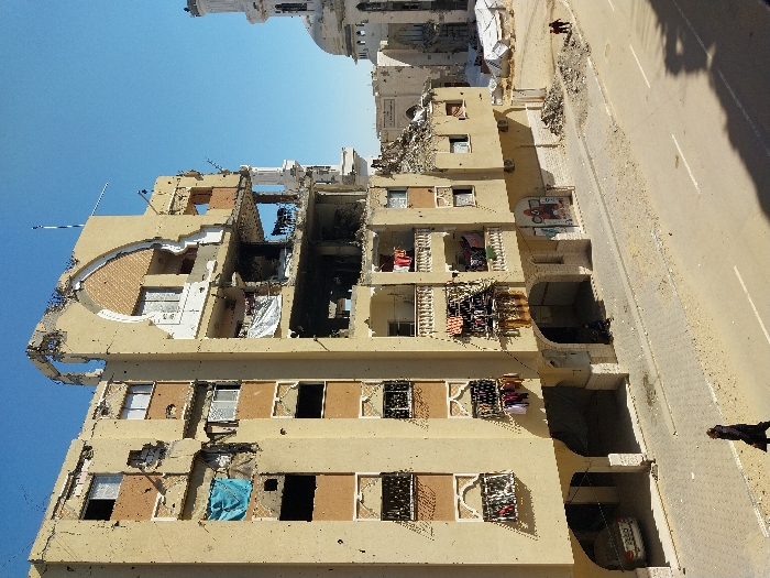 The image depicts a street scene in Khan Yunis, Palestinian Territory, under a clear blue sky, featuring a heavily damaged multi-story residential building and a partially visible mosque.
The prominent structure is a six-story building, light beige in color, with significant structural compromise. Large sections of its facade are missing, exposing interior rooms and concrete framework, particularly on the upper floors. Extensive bullet holes or shrapnel marks are visible across the remaining exterior walls. An ornate, arched architectural element on the upper-middle facade is partially destroyed. Clothes are hanging to dry on several balconies on the lower-middle floors, indicating continued habitation despite the damage. A blue tarp covers an opening on the second floor from the top on the left, and a white sheet hangs from a damaged section on the right side of the building. The ground floor features arched openings, with a silver sedan parked under one on the far left. A colorful mural, possibly depicting "Squid Game" characters, is visible on a wall section near the ground floor.
A two-lane asphalt road with a dashed white centerline and a solid white shoulder line occupies the foreground and mid-ground. The roadside and sidewalks are partially covered in dirt and debris. One individual in dark clothing walks on the sidewalk in the foreground, moving from left to right. Another person is seated on the ground near an arched opening on the ground floor of the damaged building. Further down the street on the right, two smaller figures are walking on a dirt path next to a pile of rubble.
In the background, to the right of the damaged building, a white mosque with a large central dome and a tall, slender minaret is visible. This mosque also shows signs of wear or damage, with darkened areas on its facade. Near the mosque, several white tent-like structures are present on a cleared, sandy area. Other damaged or partially collapsed buildings are discernible behind the main structures. Sunlight casts shadows across the street in the foreground and from the buildings.