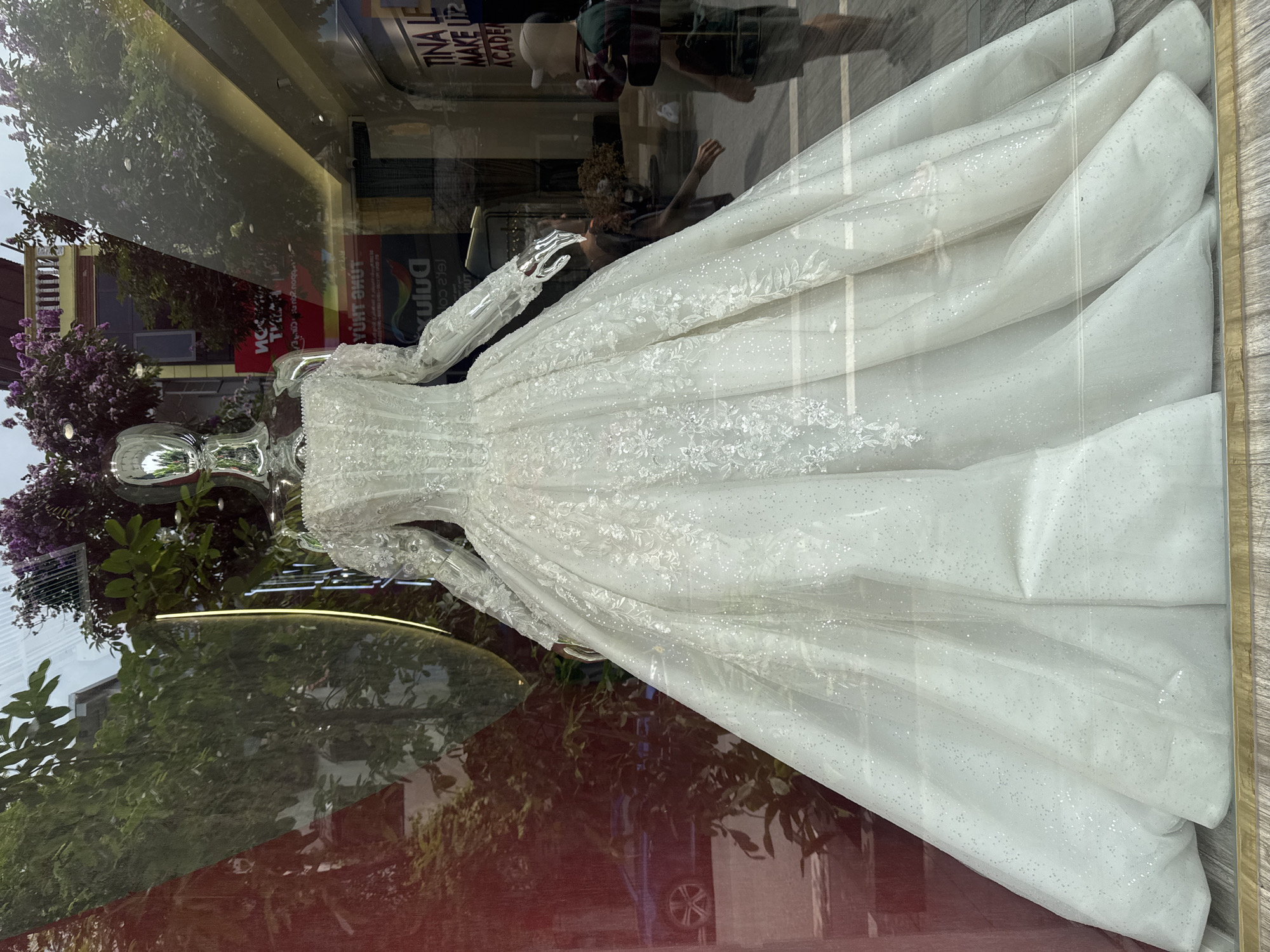 A white wedding gown on a mannequin is displayed in a storefront window. The mannequin is chrome-colored and has no discernible facial features. The dress is long-sleeved, with a fitted bodice that flares into a full skirt. The dress and the mannequin's arms are decorated with ornate floral and glitter designs.
Reflections in the glass obscure the full view. They show trees, buildings, and a person walking by the storefront. Above the dress, a sign can be partially seen reading "Tina Le Make Up". The reflections suggest that the scene is in a city, with buildings and trees lining the streets.
Based on the sign and general characteristics, the location is possibly in Hà Nội, Vietnam.