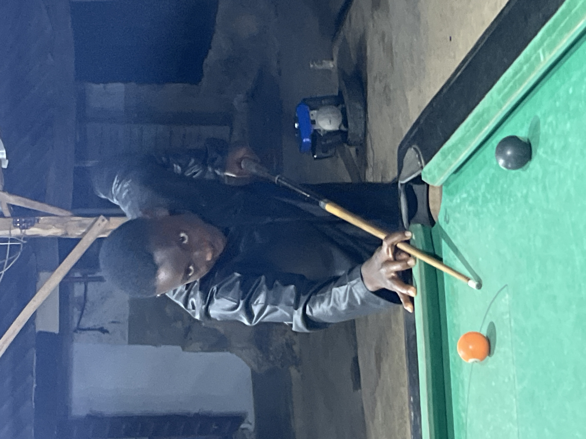 A young man is captured in a moment of intense concentration while playing pool or billiards in a dimly lit, informal setting. He is leaning over a green-felted pool table, holding a wooden cue stick with both hands, poised to strike an orange cue ball. A black ball is also visible on the table, closer to a pocket.
The man, likely of African descent, is wearing a dark, possibly leather or pleather, jacket. His short, dark hair is visible, and his gaze is fixed on the game with an expression of deep focus.
The background reveals a rustic and somewhat unfinished environment. Rough concrete walls, partially obscured by shadows, form the backdrop. To the left, wooden beams or scaffolding are visible, hinting at a utilitarian or workshop-like space. In the deeper background, on a raised surface, a small machine with blue and white components, possibly a generator or engine, is present. The low lighting suggests that the activity is taking place at night or in an interior space with minimal artificial light. There is no visible text in the image. The scene depicts a casual recreational activity in Minna, Nigeria.