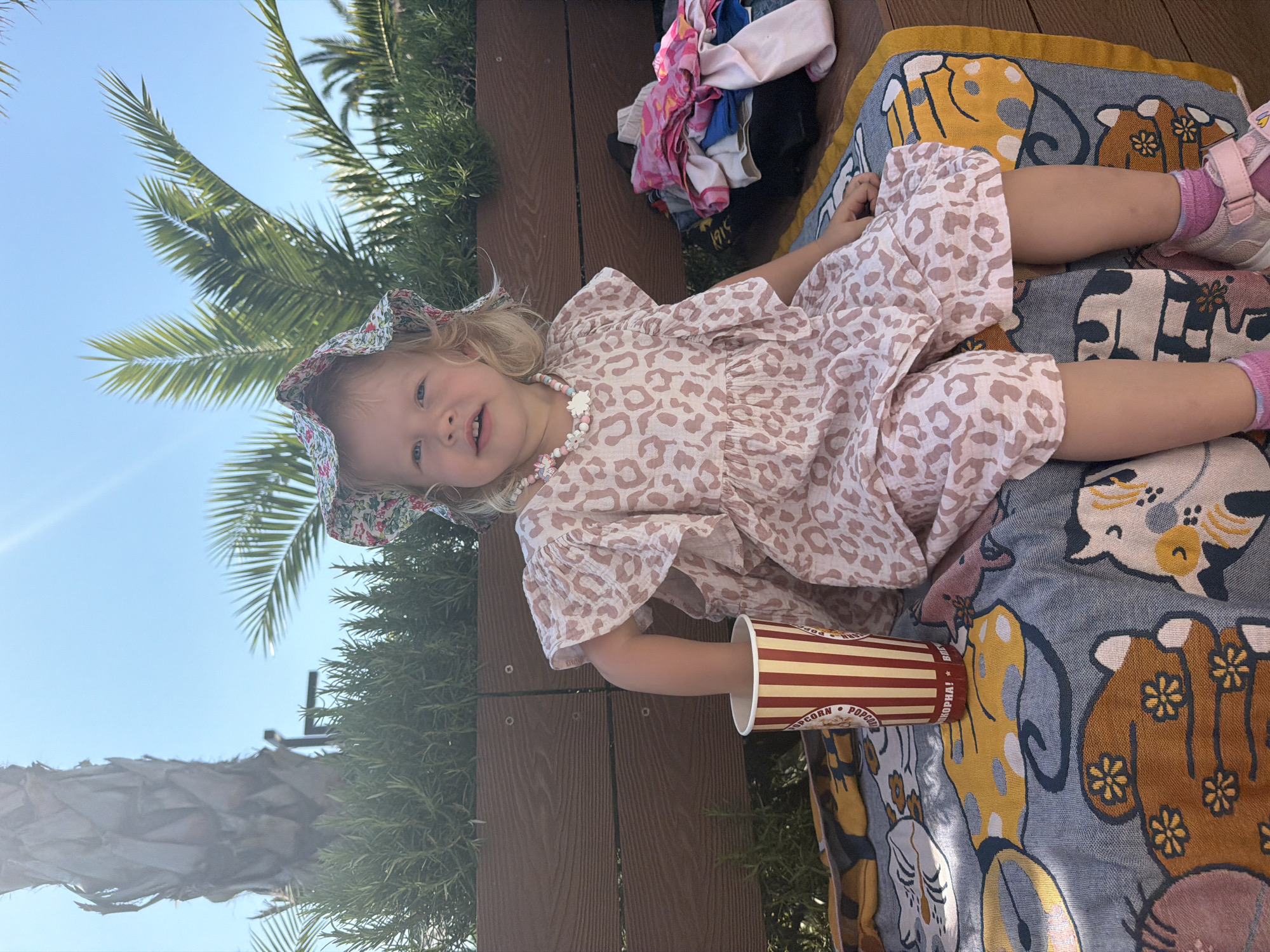 A young girl with blonde hair and a floral bucket hat is sitting on a bench outdoors. She is wearing a light pink dress with a leopard print pattern and a beaded necklace. She is holding a red and white striped popcorn cup. A colorful blanket with cartoon animal designs is spread on the bench in front of her. In the background, palm trees and greenery are visible against a clear blue sky. The setting appears to be a park or outdoor recreational area in Sochi, Russia, suggested by the tropical foliage and bright sunshine.