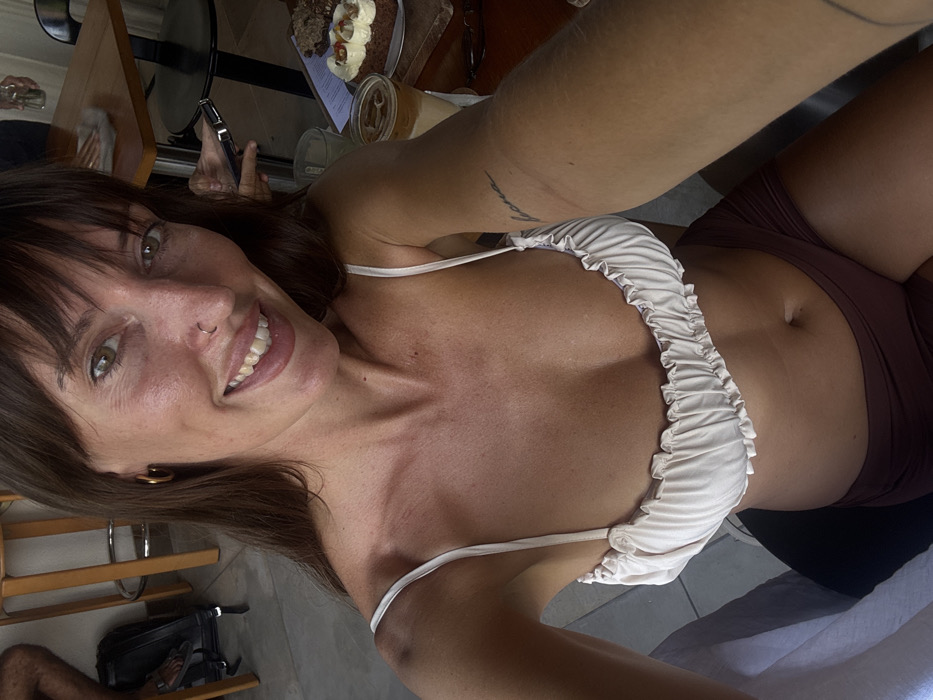 A female subject is shown from a first-person perspective, smiling directly into the camera from an indoor cafe or restaurant setting in Tibubeneng, Indonesia. She has long brown hair with bangs, light brown eyes, a ring nose piercing in her left nostril, and a gold hoop earring in her left ear. She wears a light beige ruched bikini top with thin straps and dark brown shorts. A script tattoo is present on her right inner forearm. To the left of the frame, a person's lower leg, foot in a sandal, and a black backpack are partially visible beside a light-colored bar stool. To the right, a wooden table with a metal frame displays a layered iced beverage in a glass, two desserts on a wooden board, and a menu. A hand holding a dark mobile device is visible near the table, along with a clear glass bottle. Another individual is partially visible seated at the table.