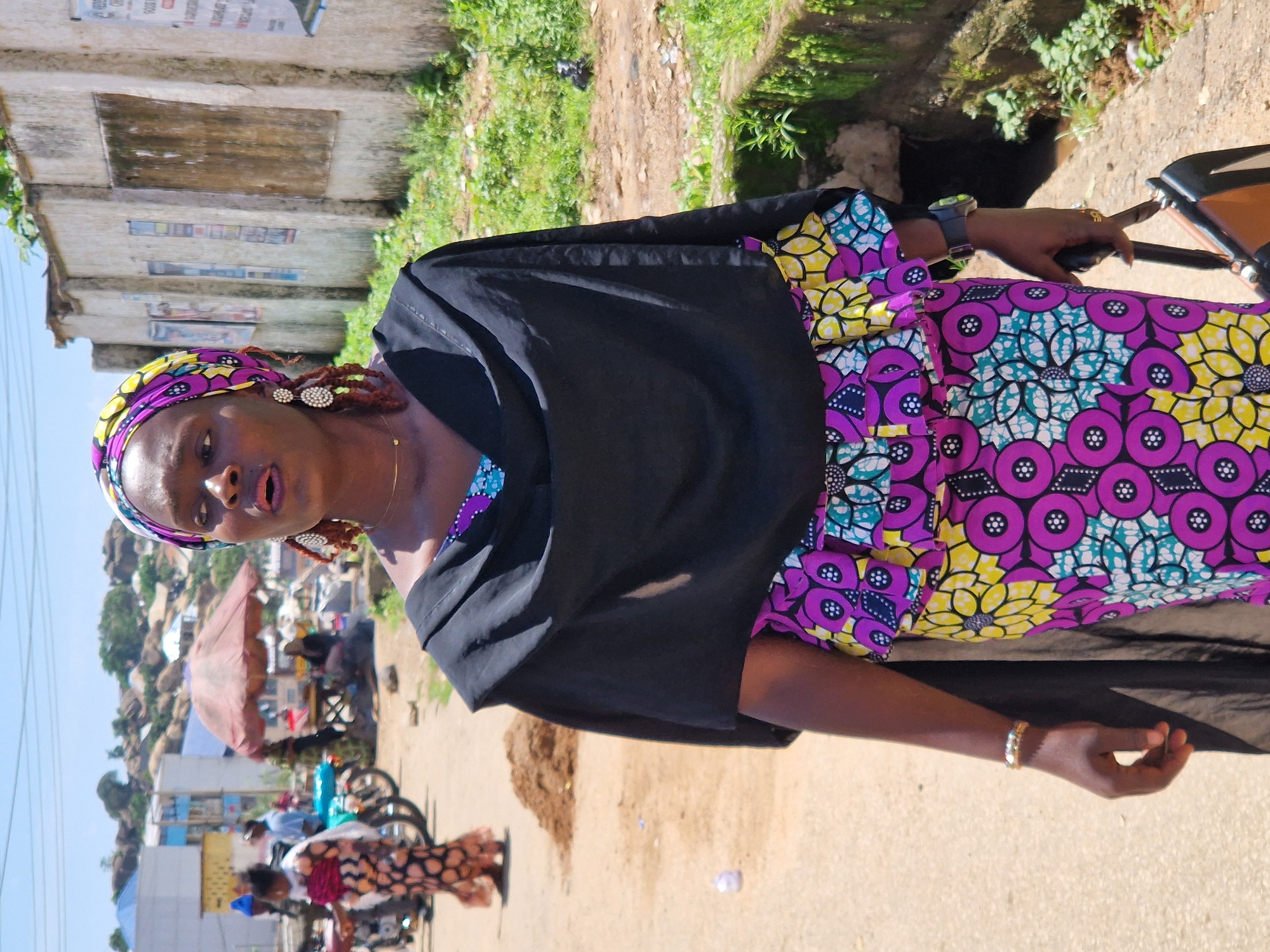 This is a medium shot photograph taken outdoors in the city of Kabong, Nigeria. The main subject is a Black woman wearing a colorful headscarf and a black shawl over a vibrantly patterned dress. The dress features a repeating floral motif in shades of purple, yellow, and blue. She is also wearing large, white, beaded earrings and a thin gold necklace. Her right hand is holding a black handbag with brown leather accents, and she is wearing a watch on her left wrist. She has a bracelet on her right arm. In the background, there is a rough, sandy path, and a wall with some posters. To the left, other individuals and motorcycles are visible, suggesting a bustling outdoor market or street scene. The sky is bright and clear, indicating a sunny day. The overall setting appears to be an urban environment with some vegetation and buildings in the distance.
