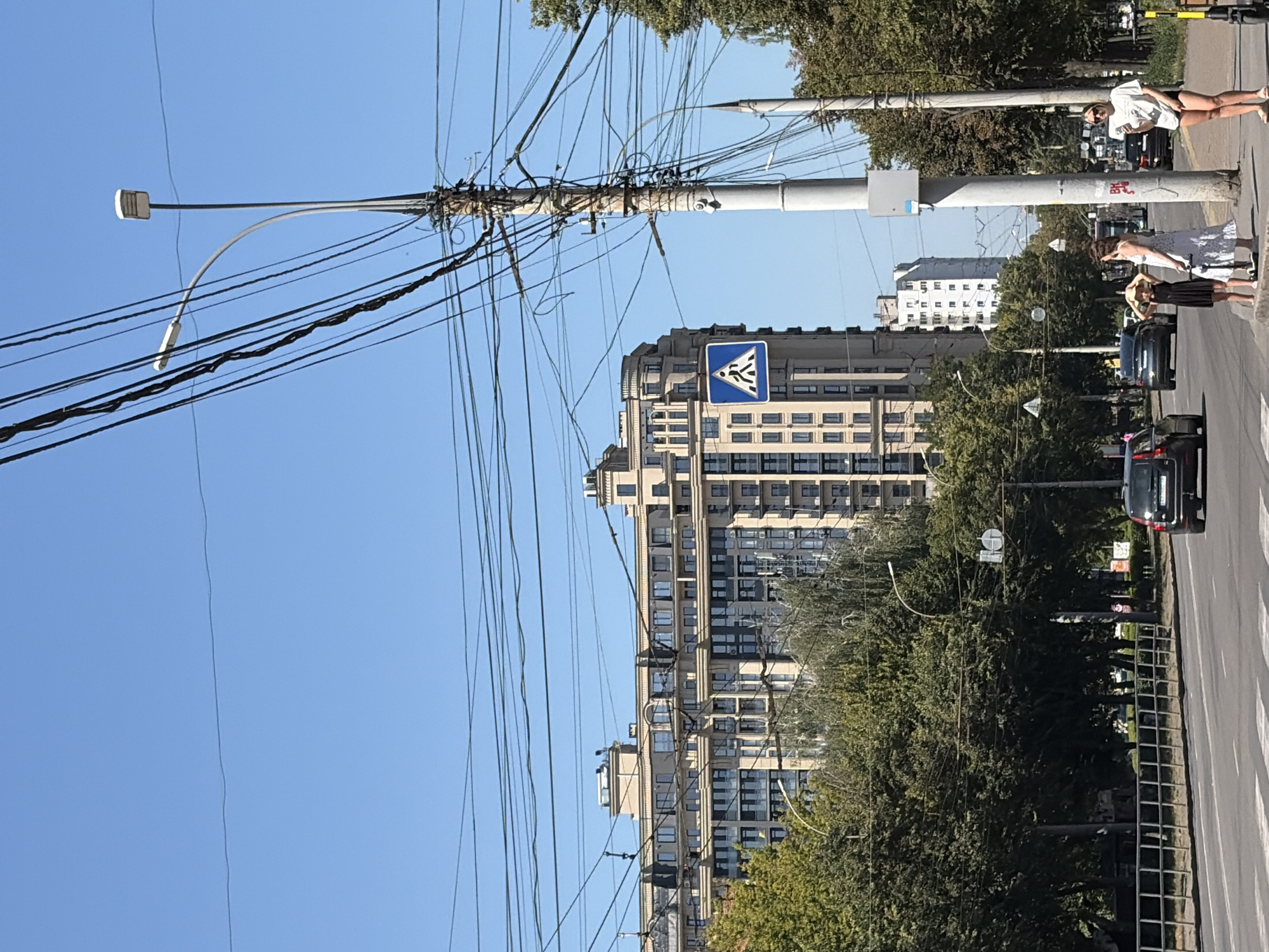 The image depicts a street scene in a city with a clear blue sky. A multi-story, modern building with a light-colored facade and numerous windows dominates the background. In the foreground, a wide road with a pedestrian crossing is visible. Several cars are on the road, and a few people are standing on the sidewalk. A utility pole with many wires is a prominent feature on the right side of the image, alongside a street lamp and a pedestrian crossing sign. Trees line the street, and another tall building can be seen in the distance. The lighting suggests it is daytime.