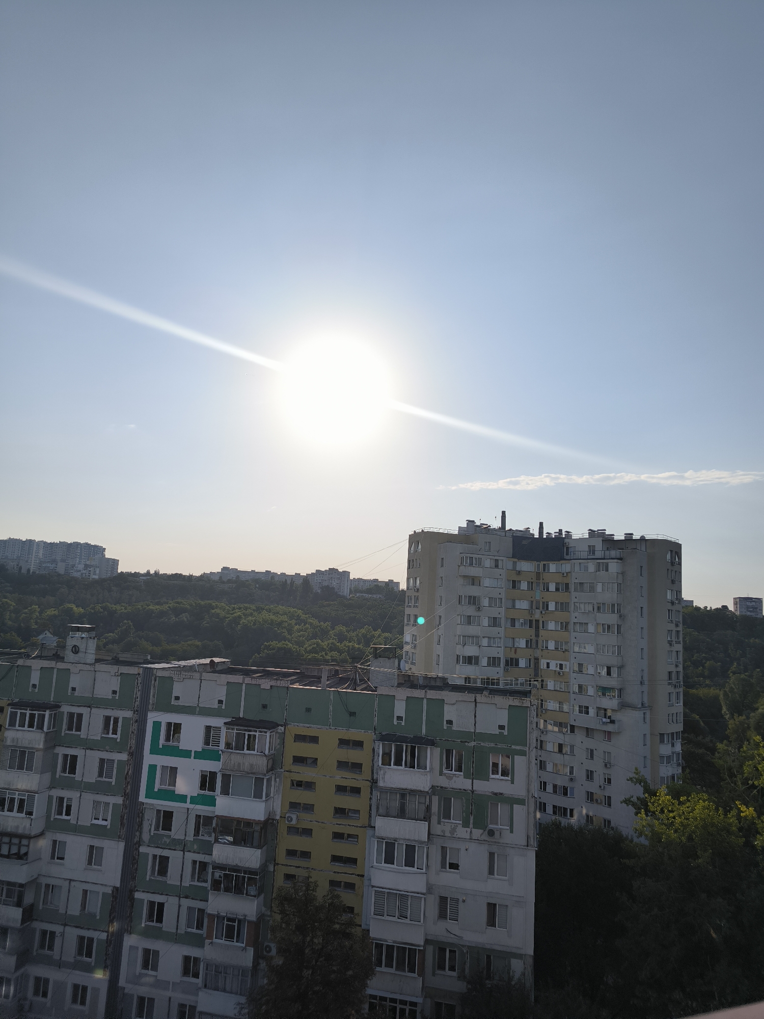 This is an aerial, daytime, exterior, eye-level, medium shot of an urban landscape in Chișinău, Moldova. The scene features several Soviet-era apartment buildings and a lush, green forest. The sun is bright and positioned in the upper left quadrant of the sky, casting a lens flare across the image. The sky is a clear, light blue with a few wispy clouds.
The apartment buildings are of varying heights and architectural styles, characteristic of the late Soviet period. The building closest to the foreground is a multi-story structure with a light green and white facade. The next building to the right is taller and predominantly yellow and gray, with visible air conditioning units on its exterior. Further in the background, more apartment buildings are visible, receding into the distance. The forest appears dense and covers the hills surrounding the city.
There are no people visible in the image. The scene depicts a quiet, residential area. The overall impression is one of a warm, sunny day in a built-up area with significant natural greenery. The photograph seems to be taken from a high vantage point, possibly a window or balcony of another building.