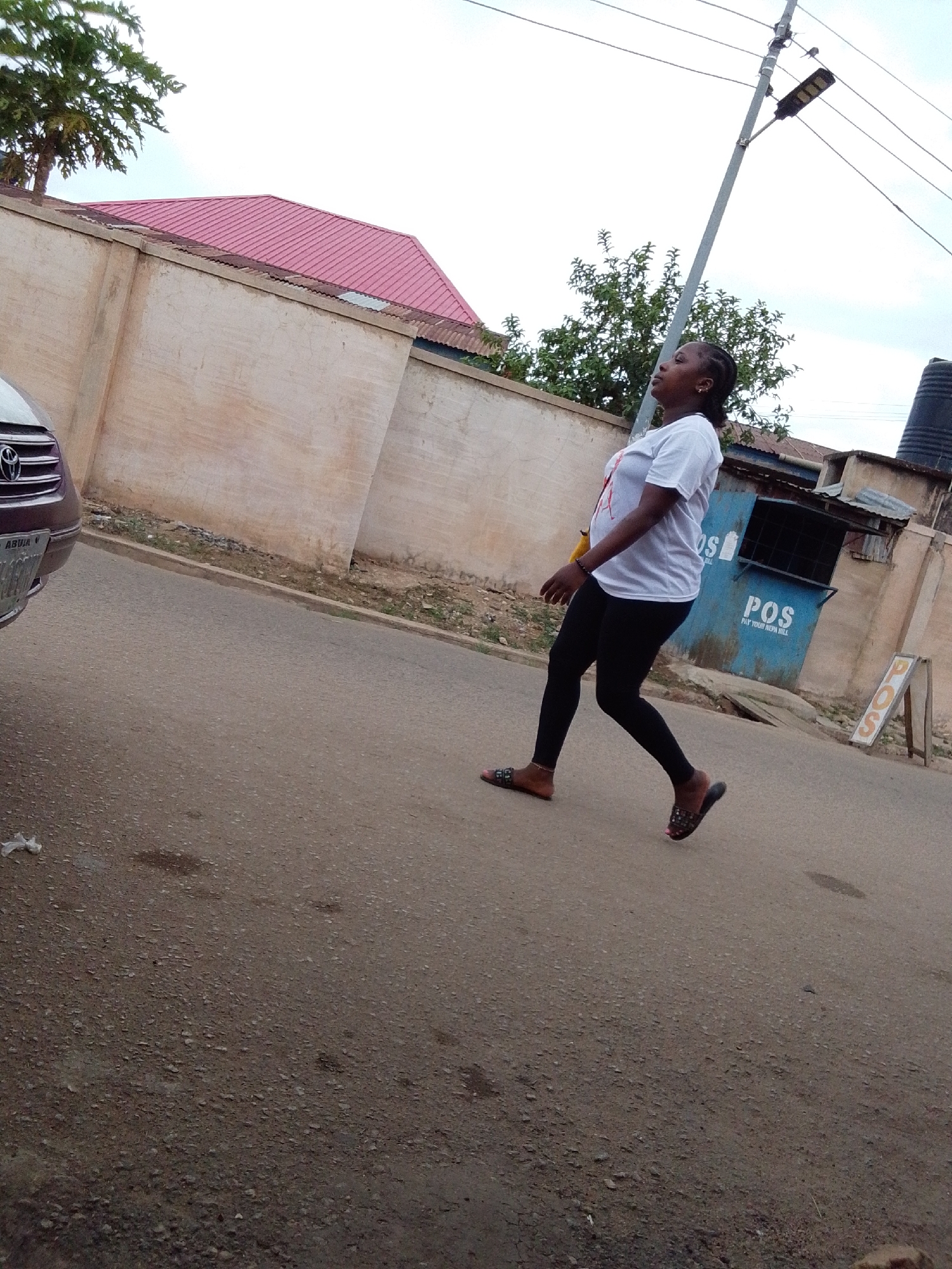 A daytime outdoor scene depicts a woman walking on a paved road. She is positioned towards the center-right of the frame, moving from the viewer's left to right. The woman wears a white short-sleeved t-shirt, black leggings, and patterned slip-on sandals. A small yellow bag or pouch hangs from her shoulder. Her head is slightly tilted upwards, and she appears to be looking towards the sky or an object overhead.
To the left, the front part of a silver or grey car is visible, featuring a Toyota emblem on its grill. Its license plate clearly shows "ABUJA" and partial alphanumeric characters. The road surface is asphalt, showing some wear and scattered debris.
In the background, a tall, light-colored concrete wall runs across the mid-ground. Above the wall, a building with a vibrant red corrugated metal roof is visible, along with green trees. Further to the right, a blue kiosk or stand with "POS" written in large white letters is prominent. Below "POS," the text "PAY YOUR NEPA BILL" is also visible. An A-frame sign next to the kiosk also displays "POS." A tall utility pole with a street light extends upwards on the right side of the image. The sky is bright, suggesting an overcast or partly cloudy day, with diffused light and soft shadows.