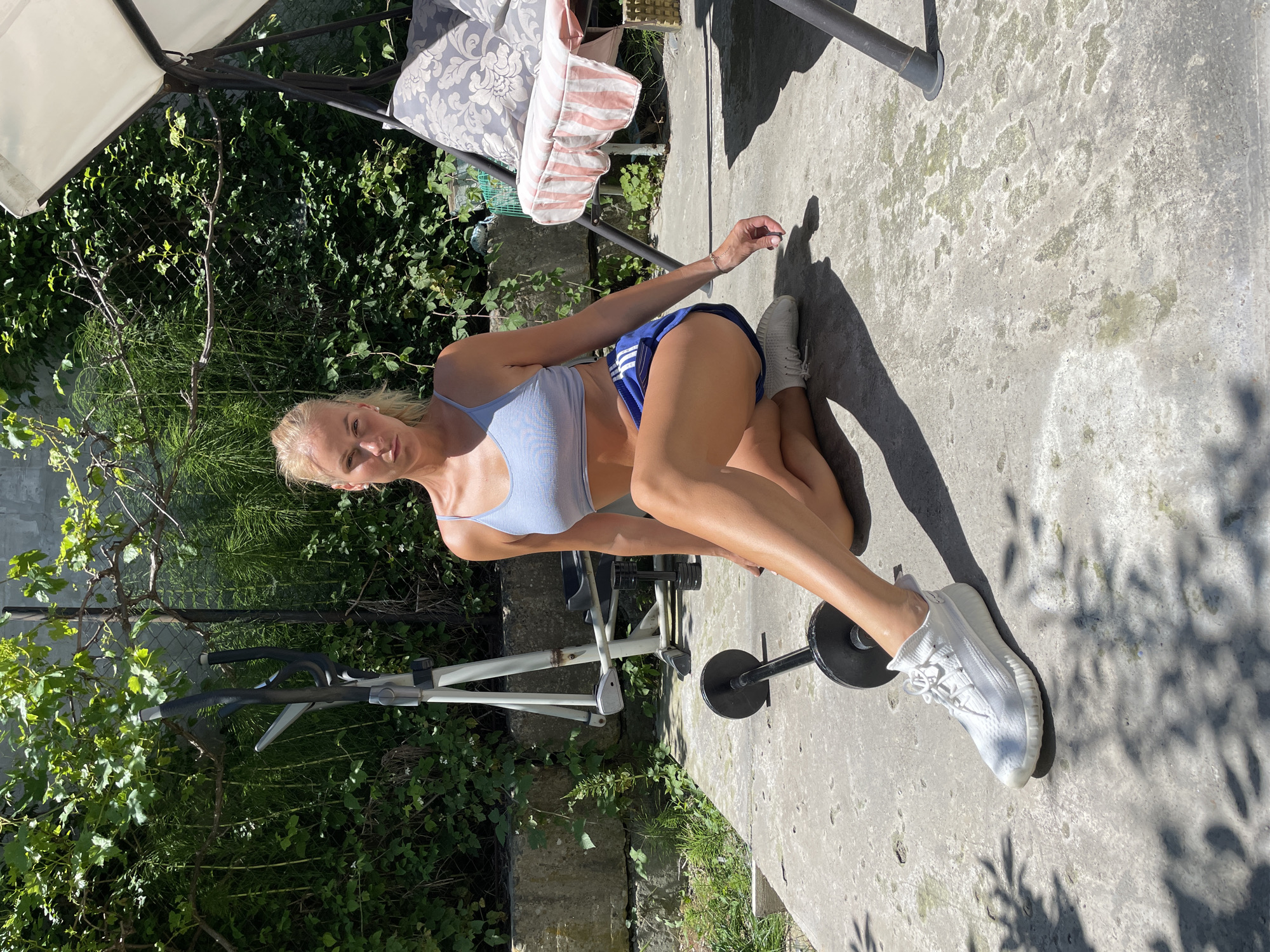 The image depicts an outdoor scene, asserted to be in Baranovka, Russia, under bright daylight. A light-skinned female, appearing to be in her 20s or 30s, is seated on a concrete surface. She has blonde hair pulled back in a ponytail and is wearing a light blue ribbed sports bra and dark blue athletic shorts with white stripes along the sides. White athletic shoes are on her feet. Her right leg is bent with the foot positioned near her left inner thigh, while her left leg is extended forward and slightly to the left. Her right arm extends behind her, with her hand resting on the concrete, and her left hand is placed on her left thigh. She looks directly at the camera with a neutral expression.
On the concrete surface near her left leg, a black barbell with black weight plates is positioned. In the background, to the left of the subject, an elliptical trainer with grey and black components is visible. Further to the right, partially visible behind the subject, is a swing chair with a white fabric canopy and cushions featuring a grey floral pattern; a pink and white striped fabric is draped over the front of its seat. The concrete ground exhibits various greenish and whitish stains and discolorations. The background is filled with dense green foliage, including bushes, vines, and taller plants, with sections of a wire fence discernible through the greenery. Strong sunlight casts distinct shadows of the subject and nearby foliage onto the concrete.