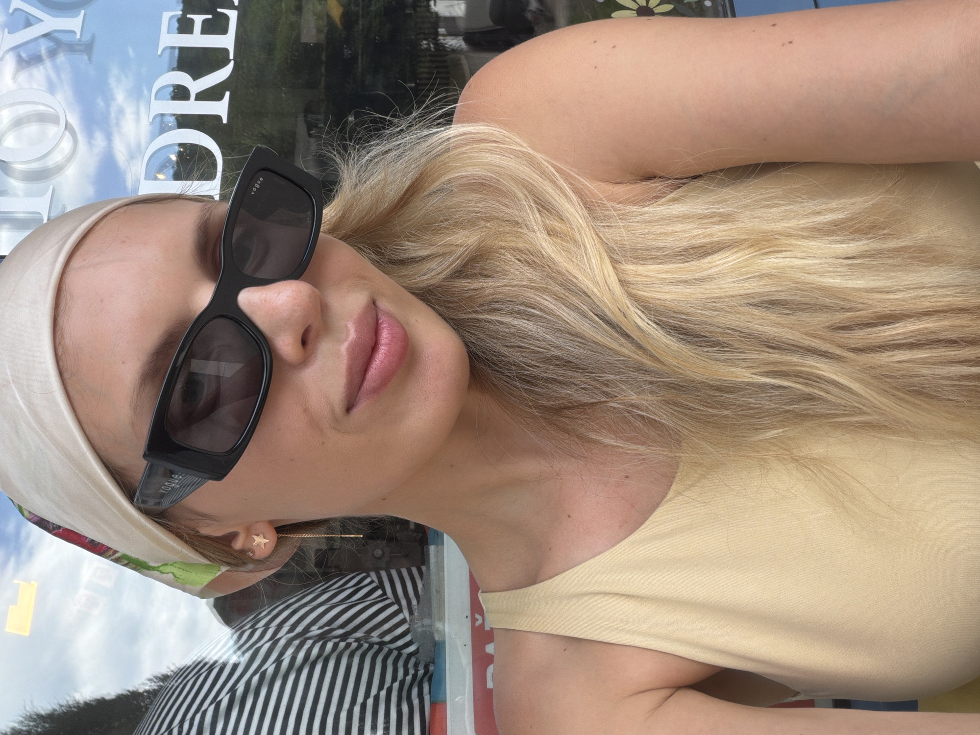 In this medium shot, a woman with blonde hair, wearing a cream-colored headscarf, black sunglasses, and a light yellow tank top, poses for a selfie. Her sunglasses feature a "Vogue" logo. She has a delicate gold earring shaped like a star with a dangling chain. In the background, through a window, a striped shirt is visible, along with text that appears to read "DREAM" on a building. The setting is likely an outdoor cafe or restaurant in Budva, Montenegro, given the style of the text and the general ambiance. The lighting suggests it is daytime.