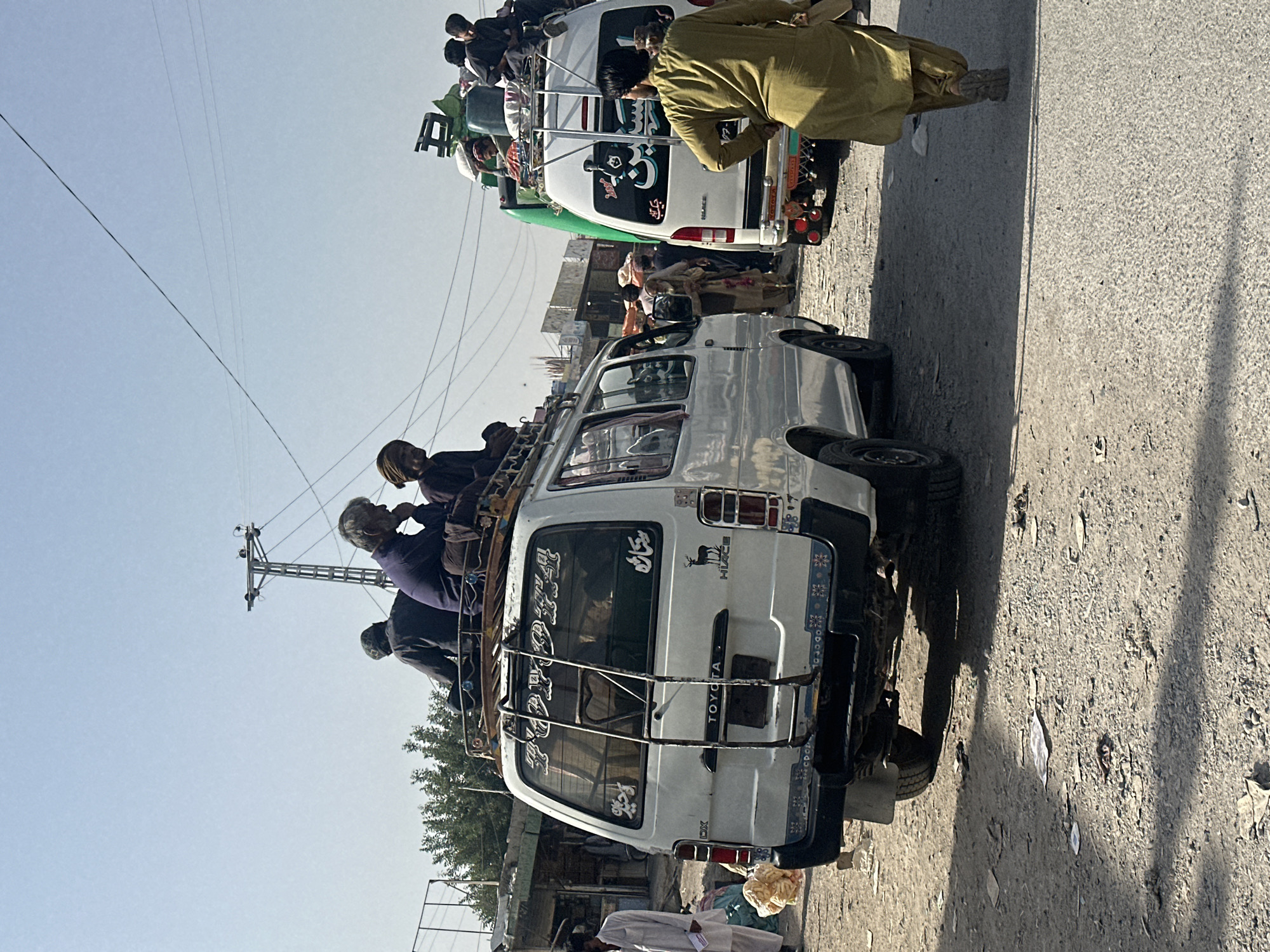 A horizontal shot shows two overloaded vans on a street in Quetta City Tehsil, Pakistan, on a sunny day.
The closer van, white, has a ladder on the back and a metal rack on top. Several people are sitting on the roof. The van has "Toyota" written on the back. A small sign decorates the upper-right corner of the window.
Behind it, a white and green van is also overloaded with people on the roof. A man in a yellow robe stands next to the vehicle.
The street is dusty and littered with debris. Power lines stretch across the sky. Buildings and trees are visible in the background.
