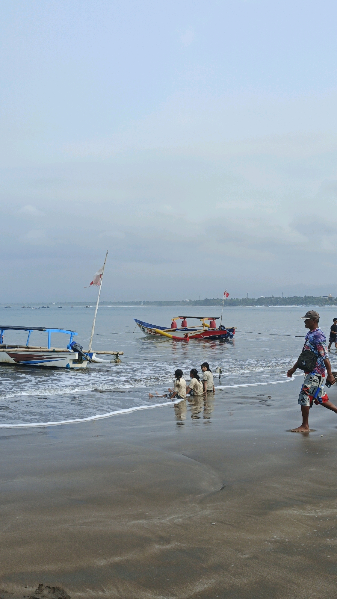 This image depicts a beach scene in Desa Pagandaran, Indonesia, under an overcast sky. The foreground is dominated by wet sand, with gentle waves lapping at the shore. In the shallow water, three young children, wearing light-colored t-shirts and shorts, are sitting on the sand, appearing to play. To their right, a man walks along the beach, wearing a patterned shirt and shorts, with a bag slung over his shoulder.
Further out in the water, several traditional fishing boats are anchored. One boat, with a blue and white hull and a blue canopy, is closer to the shore. Another, larger boat with a yellow and red hull and a red canopy, is further out. This boat has a flag with red and white stripes flying from a mast. Another boat, partially visible to the left, also appears to be a fishing vessel. The horizon shows a faint line of trees and buildings, indicating the presence of land beyond the water. The overall atmosphere is calm and serene, with the overcast sky contributing to a muted color palette.