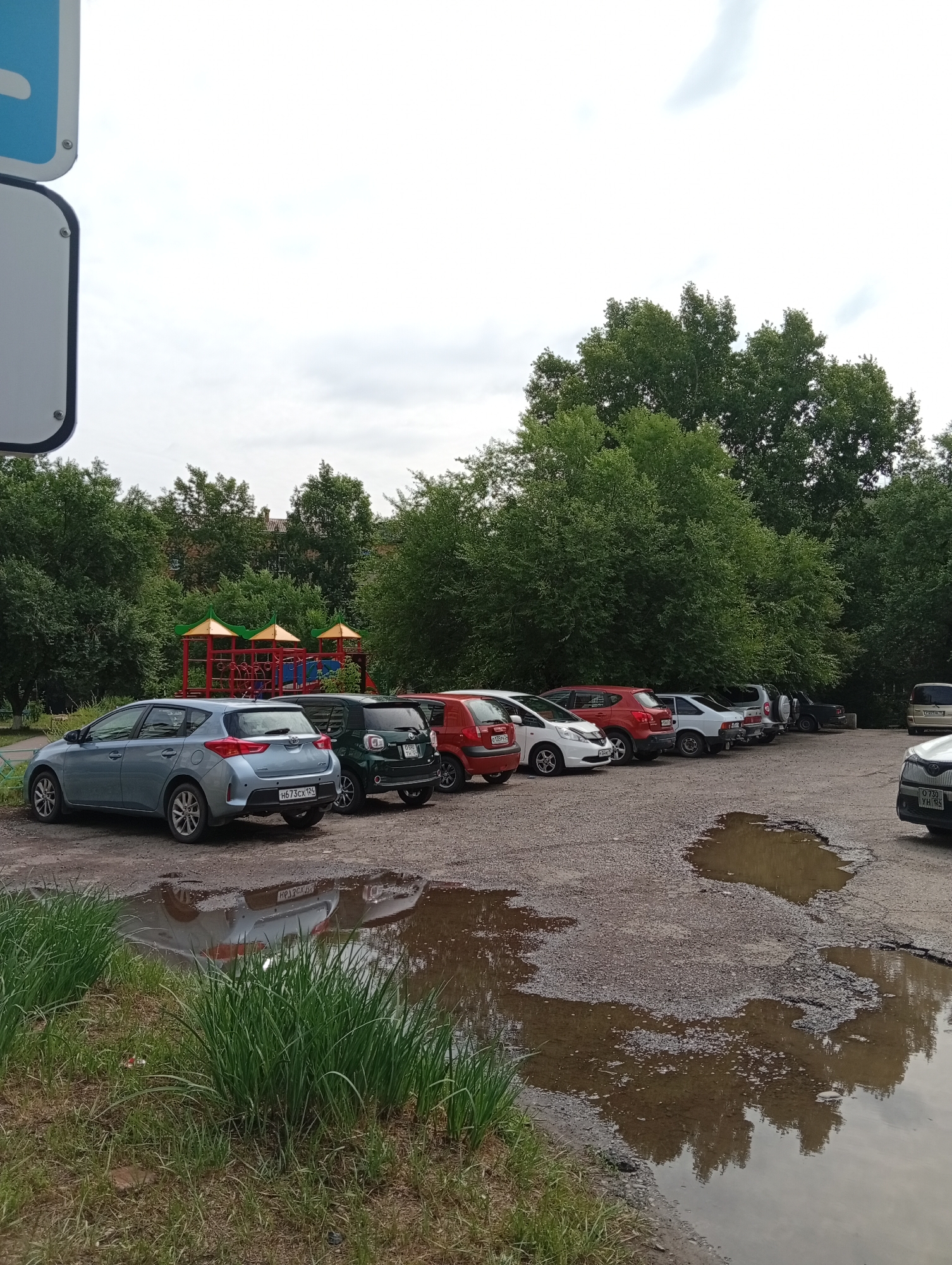 The image shows a parking lot in front of a playground with trees in the background. Several cars are parked in the lot, and there is a large puddle of water in the foreground. The sky is overcast, and the trees are green and leafy, suggesting it is summer. A sign is visible on the left side of the image, but its text is not legible. The playground equipment is colorful, with red and green structures. The overall scene appears to be a typical residential area in a city like Minusinsk, Russia, during daytime.