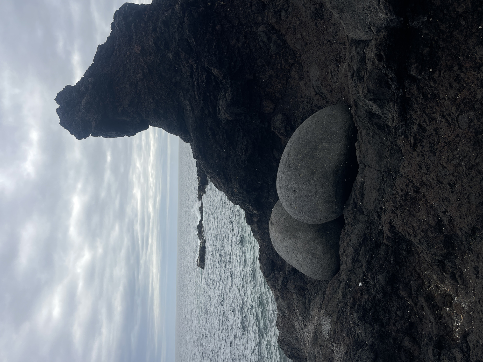 The photograph shows a coastal scene in Lagoa (Nossa Senhora do Rosário), Portugal. Two smooth, gray, round stones are lodged in a dark, rocky cliff face. Above the stones, the rock formation extends outward, forming a natural overhang. Beyond the cliff, the ocean stretches to the horizon under a cloudy sky. The water appears choppy, with a few rocky outcroppings visible in the distance.