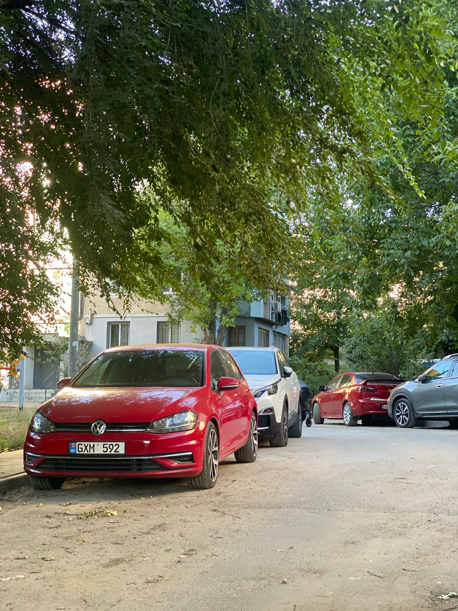 In Chișinău, Moldova, on a street bordered by trees and apartment buildings, several cars are parked. The foreground features a red Volkswagen Golf with a license plate reading "GXM 592". To its right, a white SUV is parked, followed by a red sedan and a gray SUV. The sun is setting, casting a warm light through the leaves of the trees, creating a dappled effect on the scene. The environment appears to be a residential area.