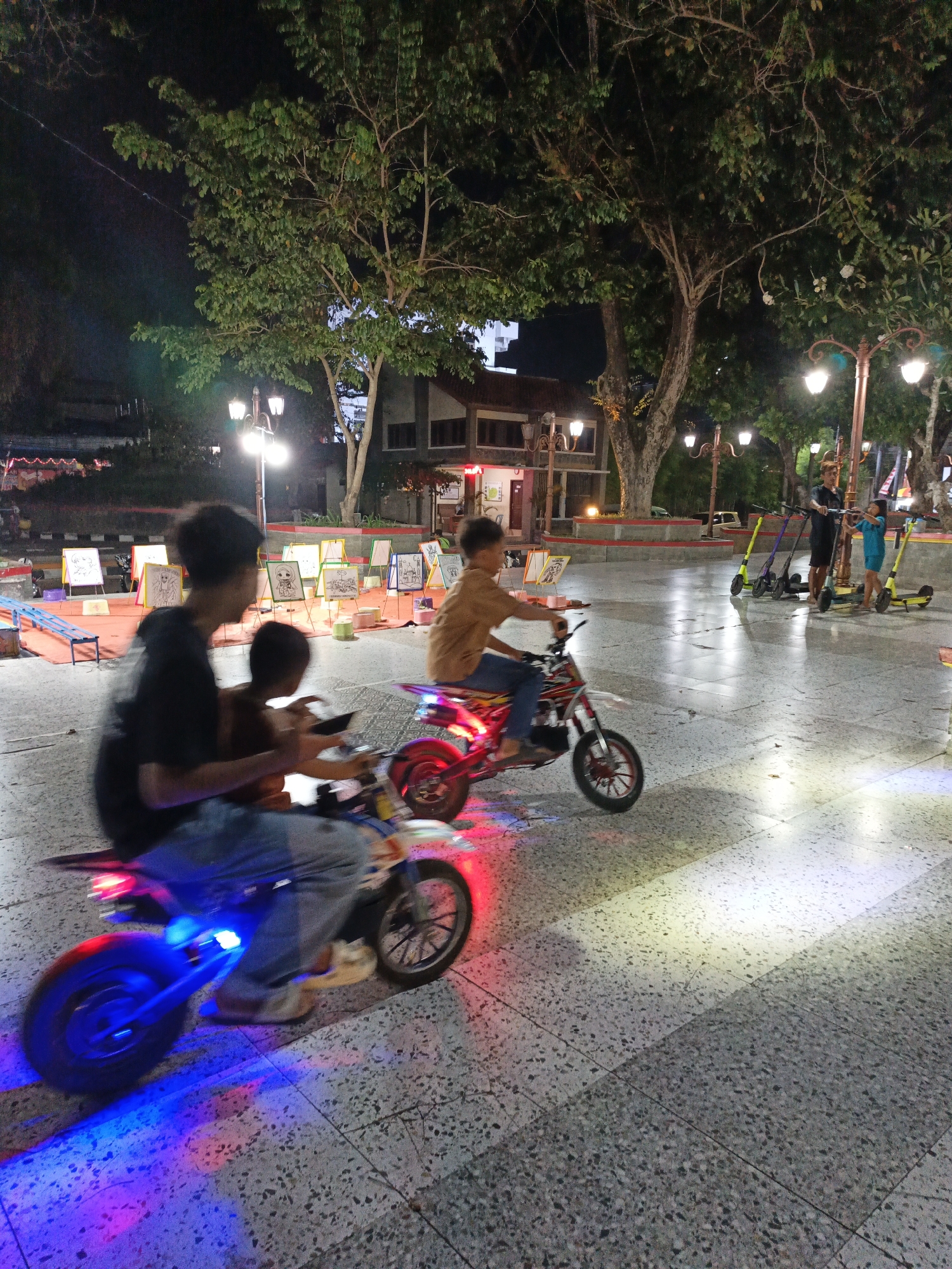 In Klaten, Indonesia, at night, children ride miniature motorcycles and scooters in a public square. The square has a patterned tiled floor and is illuminated by street lamps and the colorful lights of the vehicles. In the foreground, two boys on a motorcycle with red and blue lights are visible. The boy in the back is holding a phone. To the right, another boy is riding a red motorcycle. In the background, several other children are seen with electric scooters, and there are easels with children's drawings set up. Large trees with dense foliage are present, and buildings can be seen in the distance.