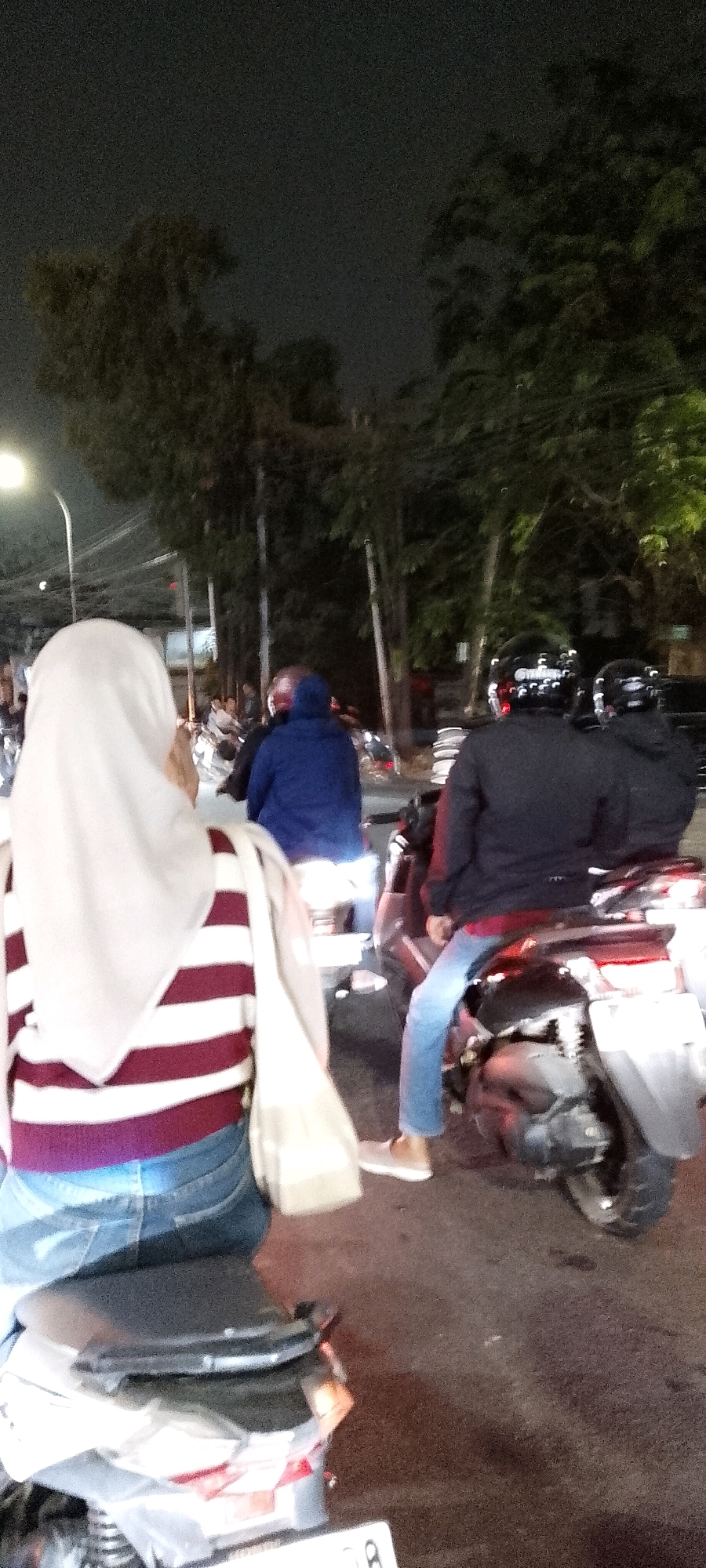 The image depicts a nighttime scene on a street, possibly in Bekasi, Indonesia, with several motorcycles and their riders. In the foreground, a person wearing a white hijab and a maroon and white striped long-sleeved shirt is seated on a motorcycle, facing away from the camera. To the right, another person wearing a dark jacket and blue jeans is riding a motorcycle, also facing away. Behind them, another motorcycle rider in a blue hooded jacket is visible. The environment appears to be an urban setting with trees lining the street and streetlights illuminating the scene. The overall atmosphere is one of everyday traffic movement at night.