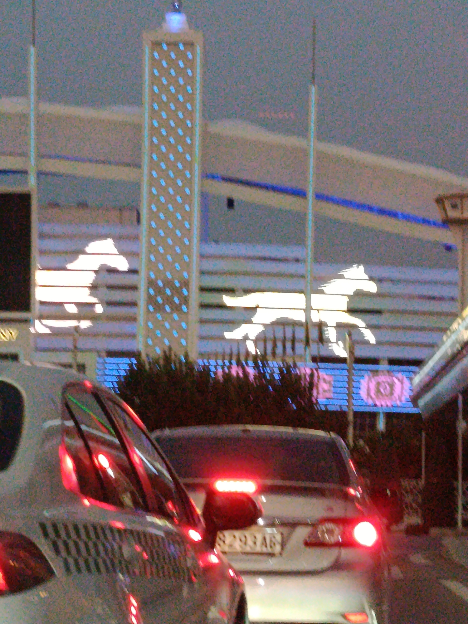In the city of Unknown, Unknown, at an outdoor location at night, a silver car is parked in the foreground. The car has its brake lights on, emitting a red glow. In the background, a tall, rectangular structure with a pattern of blue lights is visible. Silhouettes of two horses are illuminated against a striped background. A blue stripe runs horizontally across the middle ground. The sky is overcast and dark.