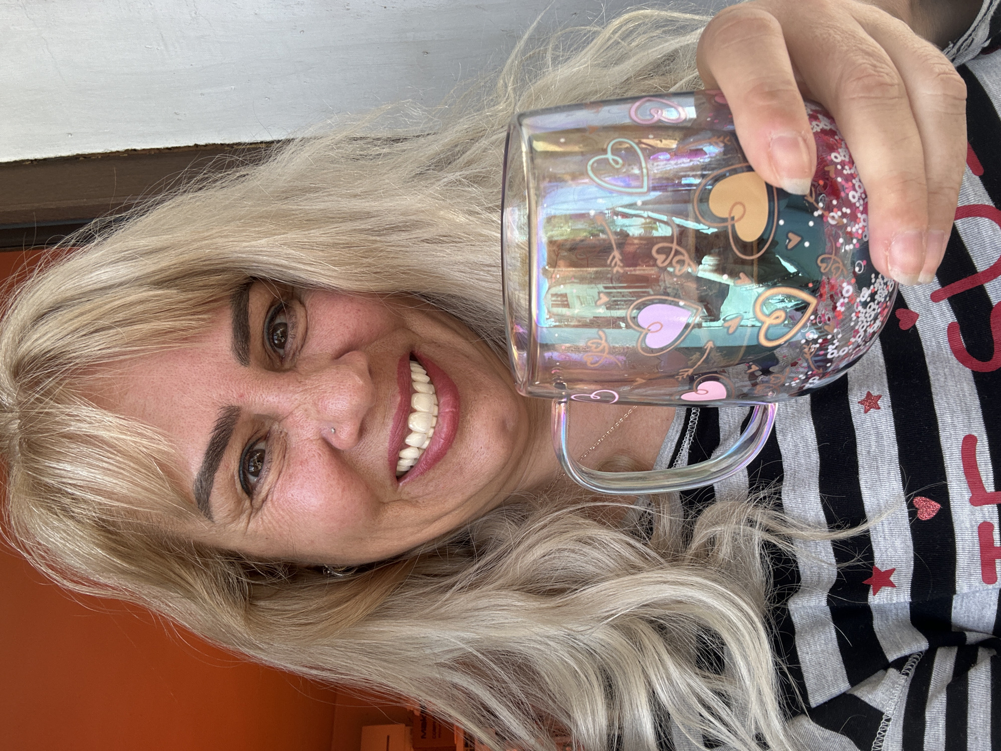 A close-up portrait shows a smiling woman holding an iridescent glass mug. The woman has long, wavy blonde hair, fair skin, and dark eyebrows, and a small piercing on her right nostril. She smiles broadly, revealing white teeth, and her dark eye is visible. She is wearing a grey and black striped top decorated with red stars and a red glitter heart. Partially visible red text on her shirt, starting with the letter "L," likely spells out "LOVE."
Her right hand grips the clear mug, which is adorned with outline hearts in pink and gold, along with arrow designs. The lower section of the mug contains a vibrant mix of red, pink, and white glitter or confetti, creating an iridescent effect. The background features an orange-brown wall or surface on the lower half, separated by a dark brown horizontal line from a lighter grey or white textured wall above. The woman's expression is joyful and cheerful. The scene is set in Sofia, Bulgaria.