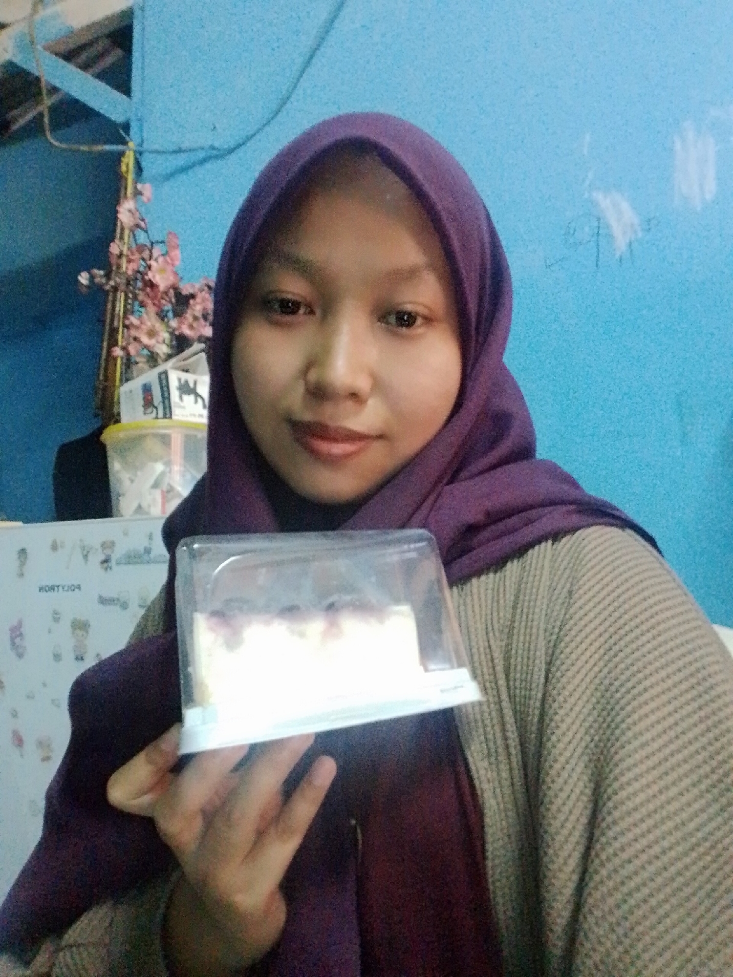 A young woman, appearing to be of Southeast Asian descent, is pictured in a close-up, shoulder-up shot, holding a clear plastic container with a slice of white cake topped with purple berries. She is wearing a dark purple hijab and a textured, light brown cardigan over a dark red top. Her skin is smooth, and she has a neutral expression. The background is a plain blue wall, with some light fixtures and a floral decoration visible on the left side. To the left of the woman, partially obscured, is a refrigerator with colorful cartoon decals. The scene appears to be indoors, likely in a residential setting. The lighting is dim, suggesting it was taken in low-light conditions, possibly indoors with artificial lighting. There are no overt indications of the specific location within Bencongan, Indonesia, beyond the general indoor setting.
