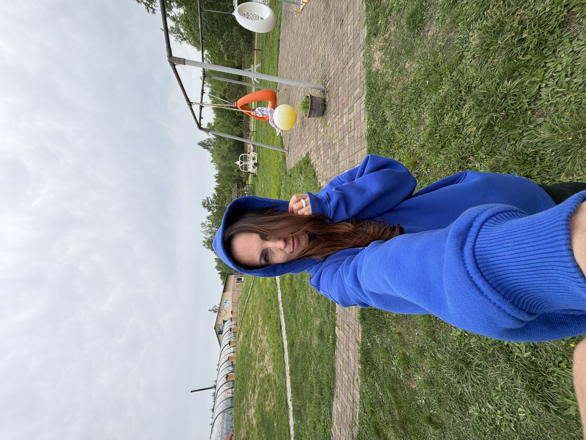A woman wearing a blue hooded sweatshirt is taking a selfie outdoors. She is standing on a grassy area next to a brick walkway. In the background, there is a playground swing set with a white tire swing, an orange swing, and a yellow ball. Further back, there is a white decorative carriage and a long greenhouse-like structure. There is also a building with graffiti on the wall. The location is Birobidzhan, Russia.