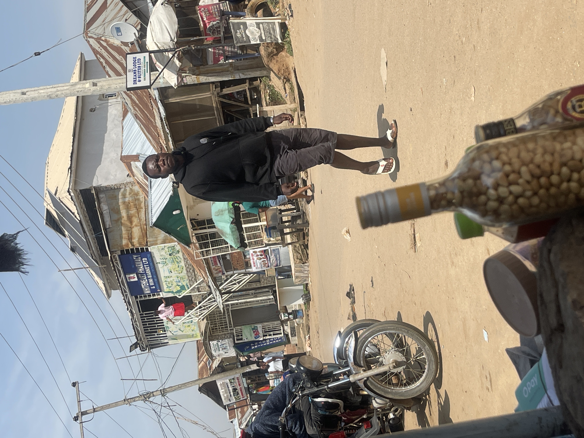 In Abuja, Nigeria, a man in a black hoodie and grey shorts walks along a dusty street lined with various shops and businesses. A motorcycle is parked on the left side of the road, with its front wheel and fender clearly visible. In the background, a woman in a pink top and red skirt stands on stairs leading up to a building. Signs indicate businesses such as "WHITEHEART PHARMACY & MINI-MARKET LTD" and "DREAMS LODGE & SUITES LTD." Several other structures, some with corrugated metal roofs, are visible. In the foreground, two glass bottles, one filled with small, light-brown nuts and another partially obscured, are positioned in front of the camera. The overall scene depicts a bustling commercial area under a bright, clear sky.